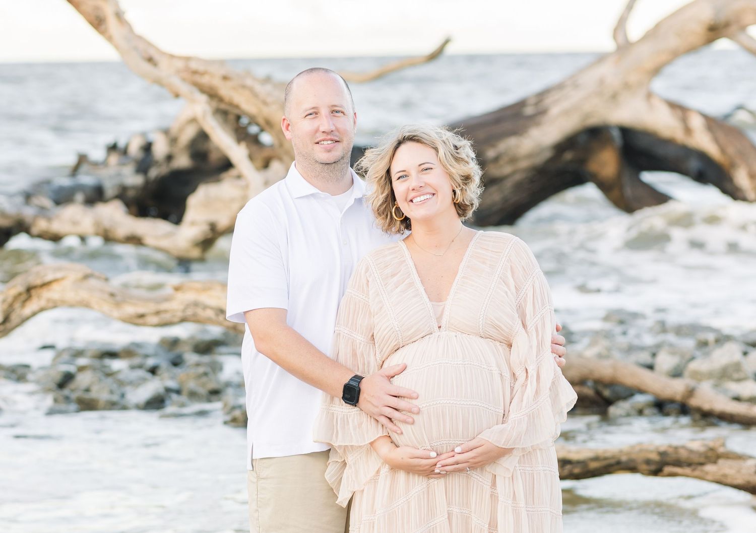 Couple in neutral tones embraces lovingly on sandy driftwood beach during sunset photoshoot.