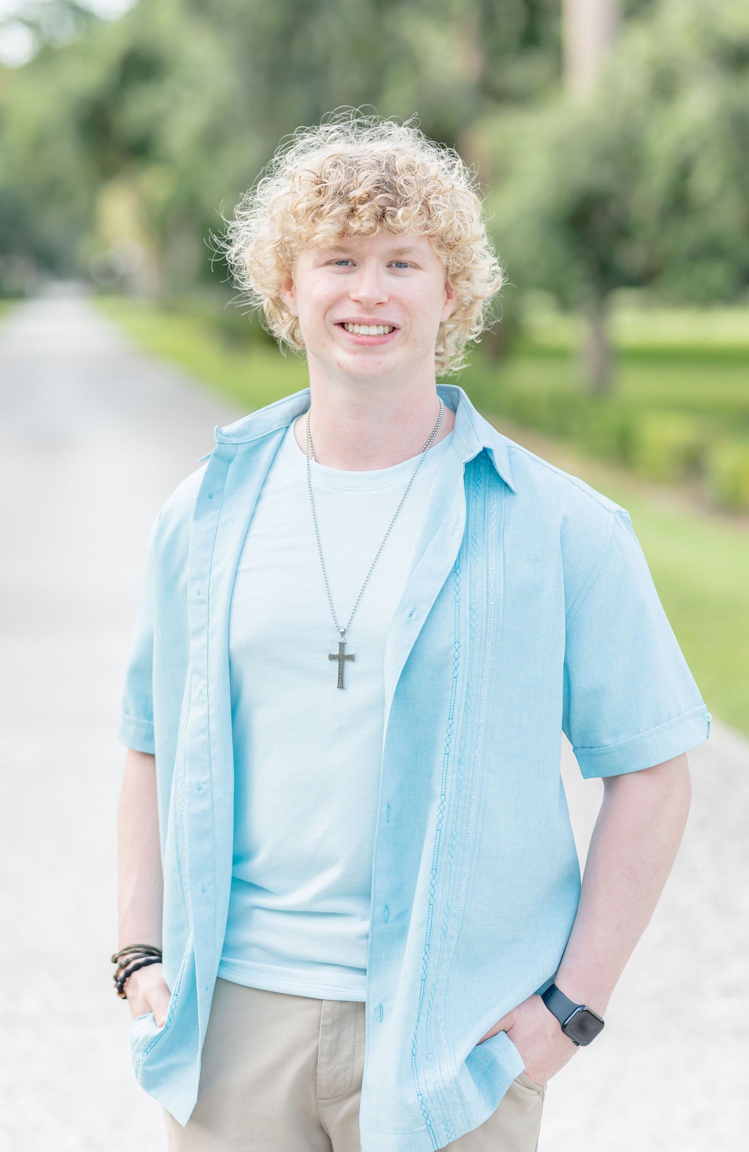 Person in light blue shirt and beige pants poses outdoors in a park setting with trees and greenery in background.