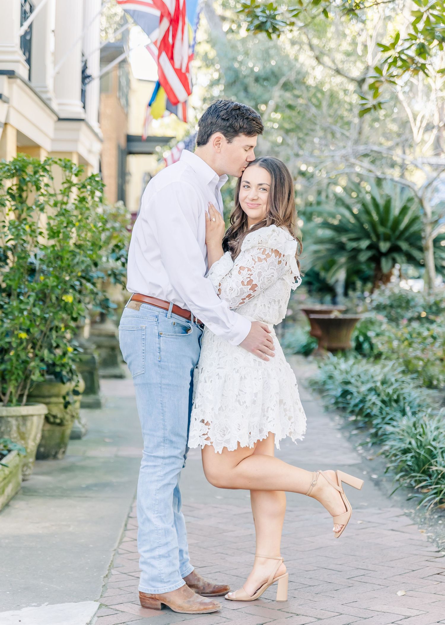 A romantic couple embraces on a brick sidewalk in Charleston, with her wearing a white lace dress and him in jeans.