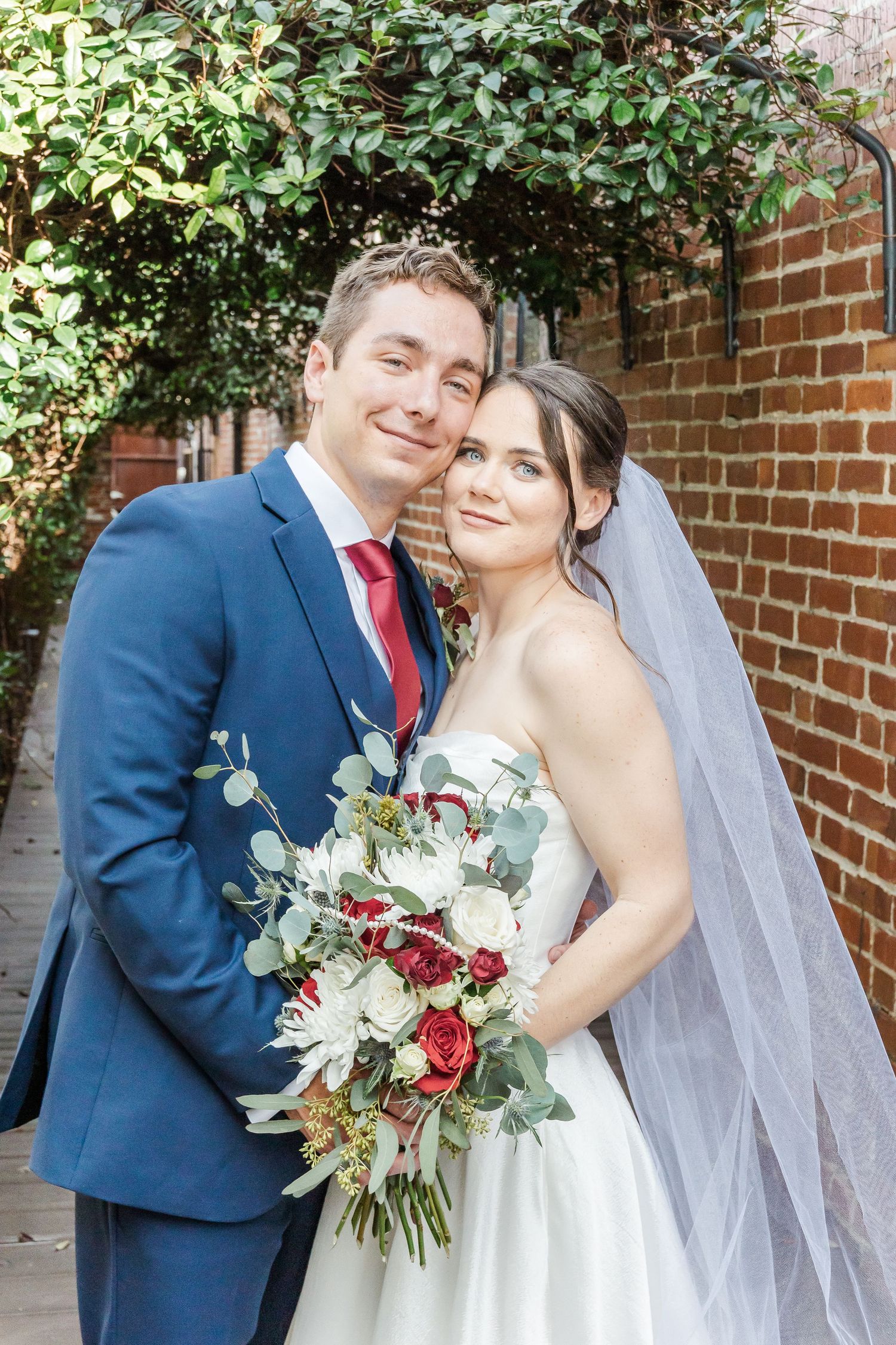 Wedding photos showing the couple embracing against a brick wall backdrop with lush greenery.