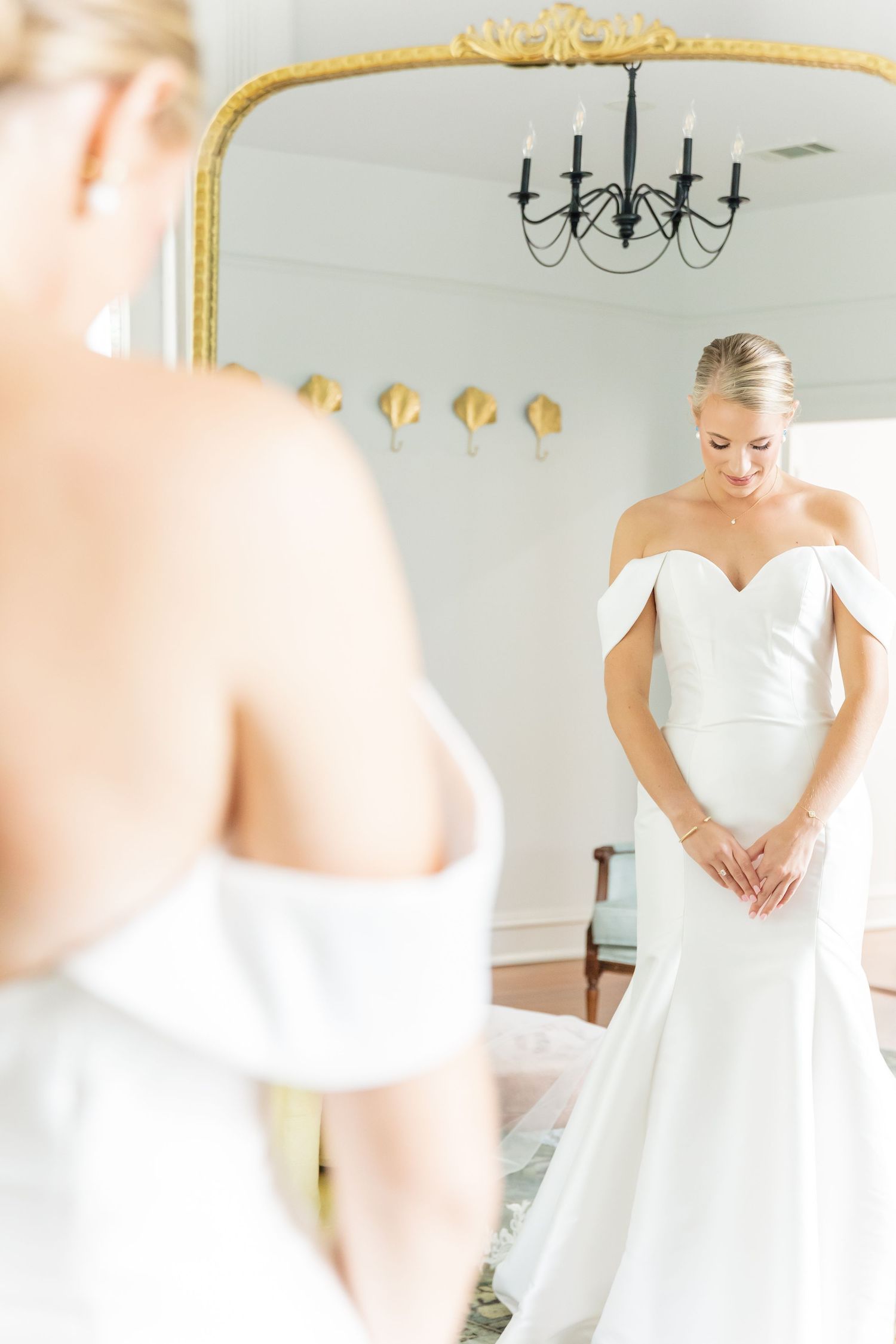 Elegant bride wearing off-shoulder white wedding gown looks down while standing in front of ornate gold mirror.
