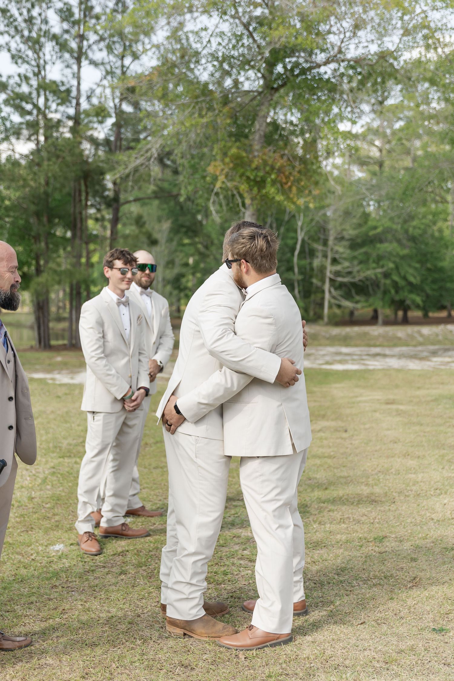 Wedding party members in beige suits gather together on grassy outdoor venue.