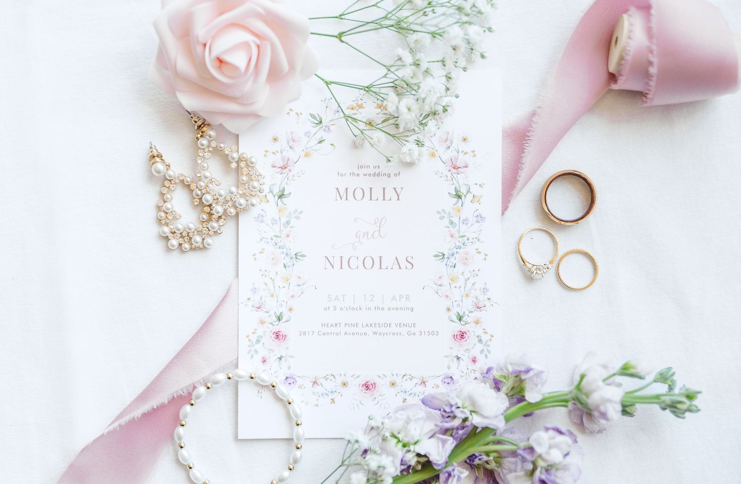 Wedding invitation flat lay with pink roses, pearl jewelry, and gold rings.