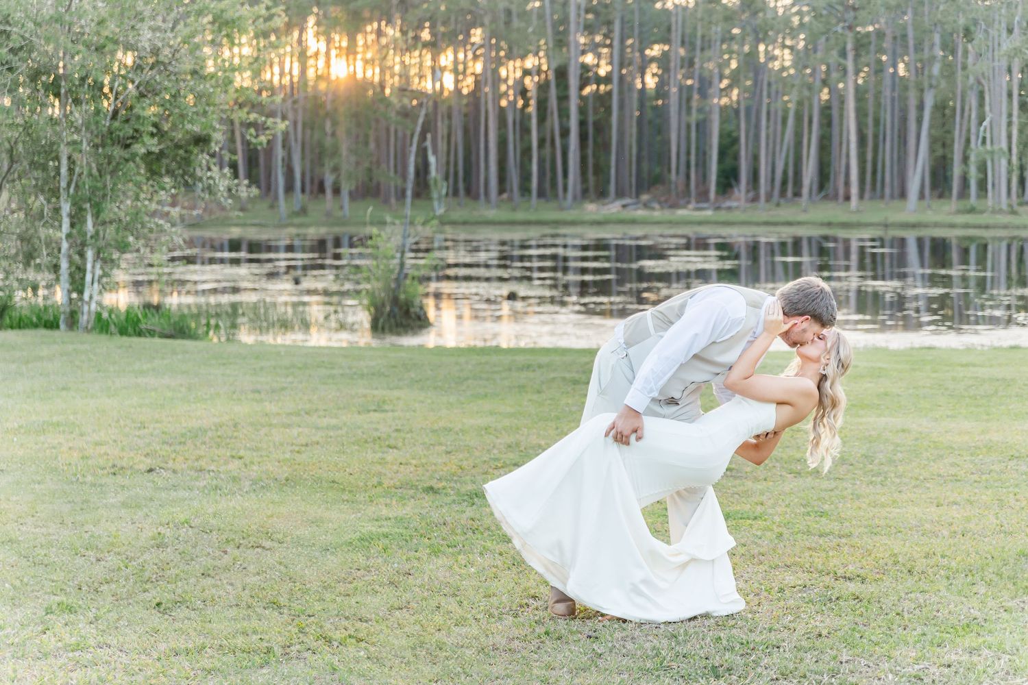 Playful wedding moment captured as couple dips dramatically on grassy lawn near lake.