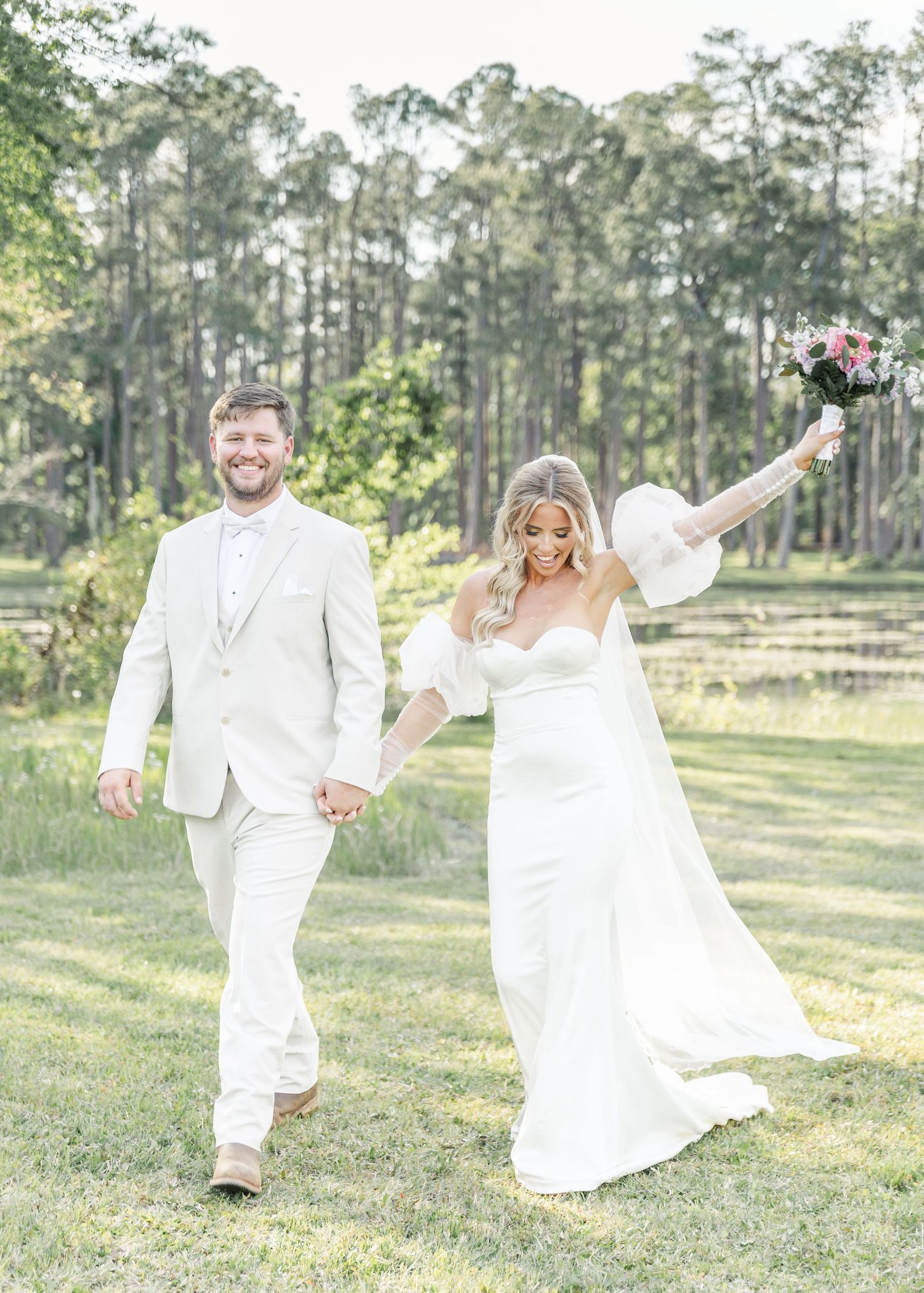 Celebratory wedding couple in white walking through a pine forest with bride holding bouquet high.