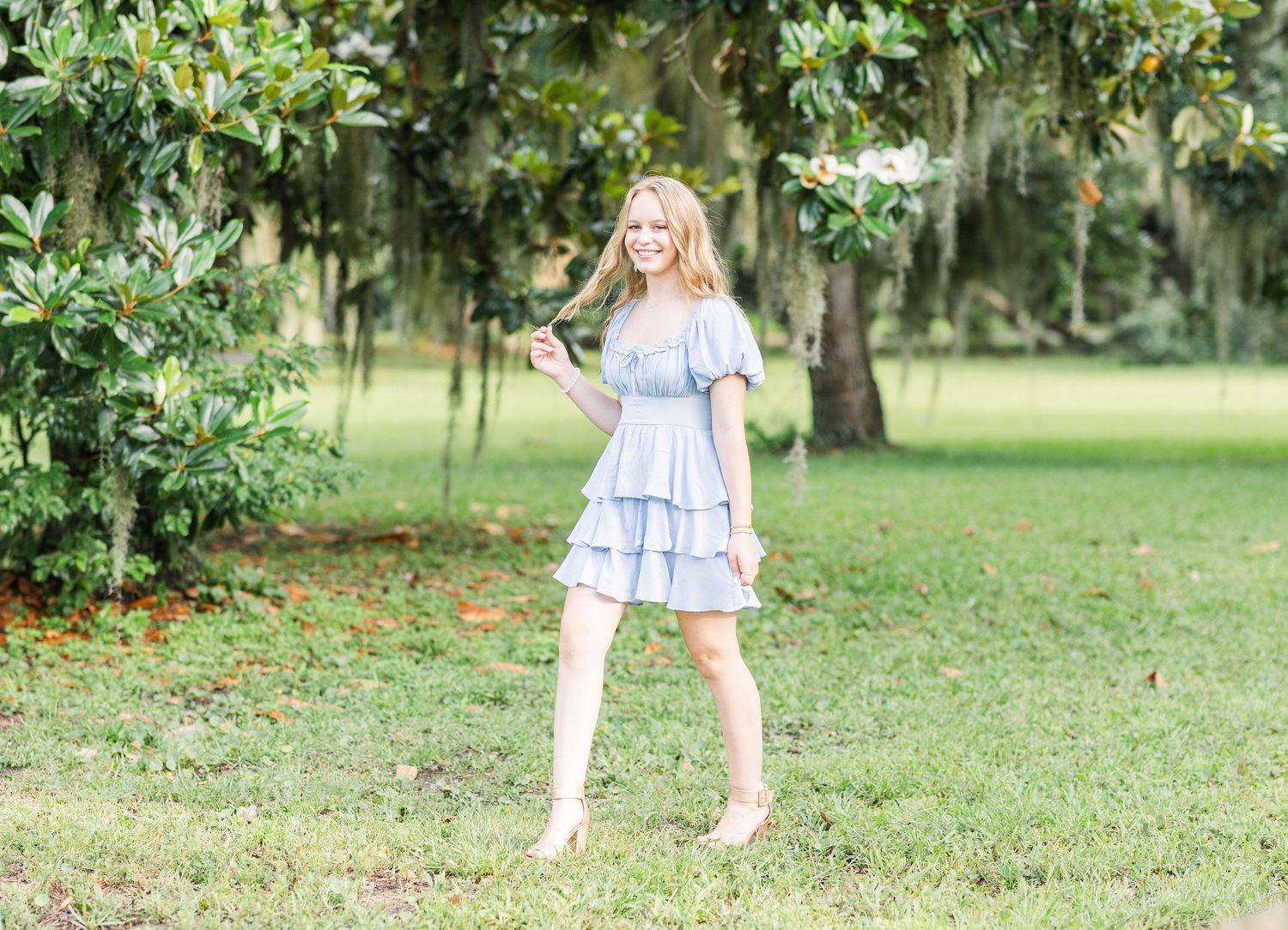 A person in a light blue ruffled dress stands on grass beneath moss-covered trees.