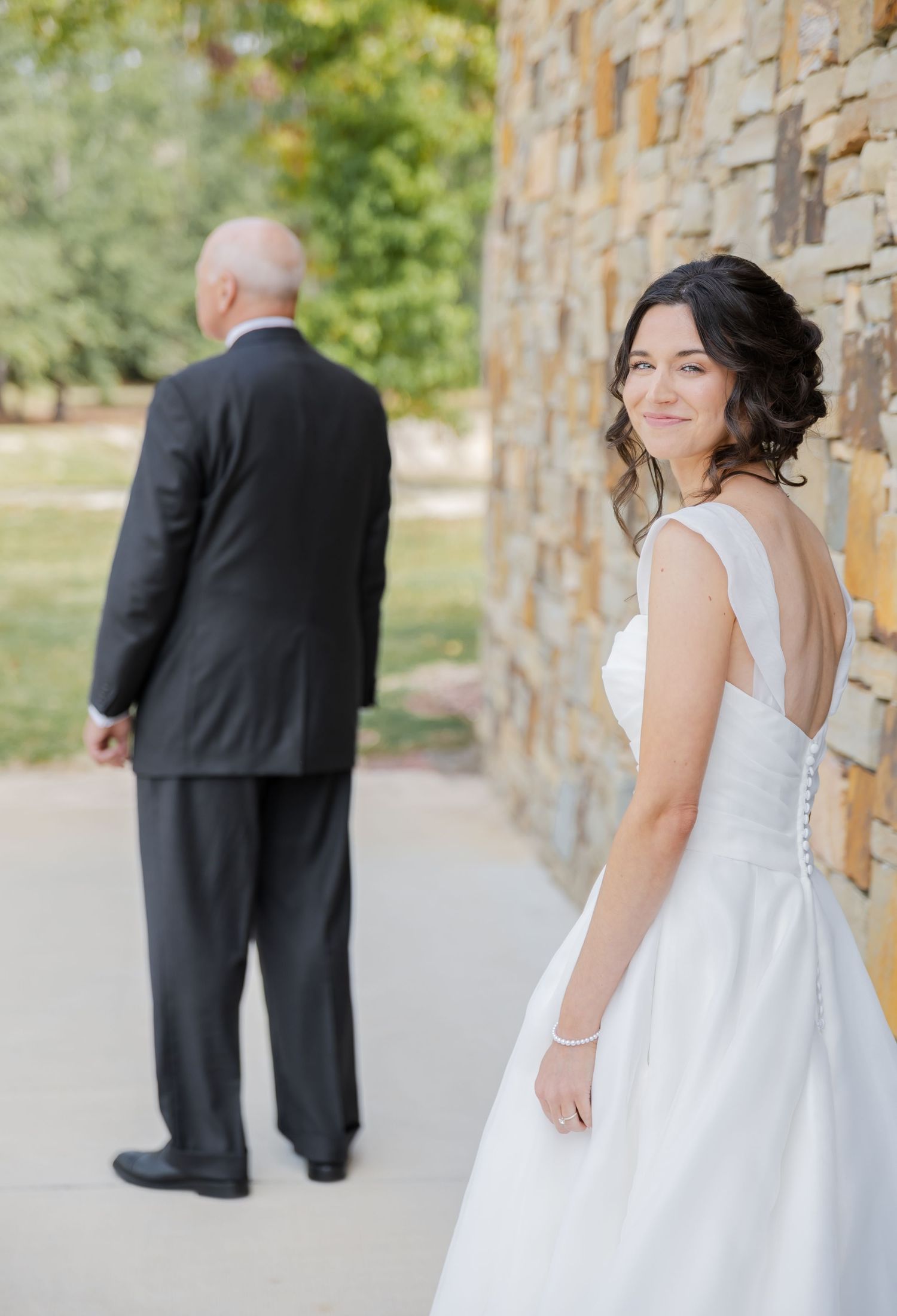 A series of wedding photos showing a first look moment between a bride in white and groom in black by a stone wall.