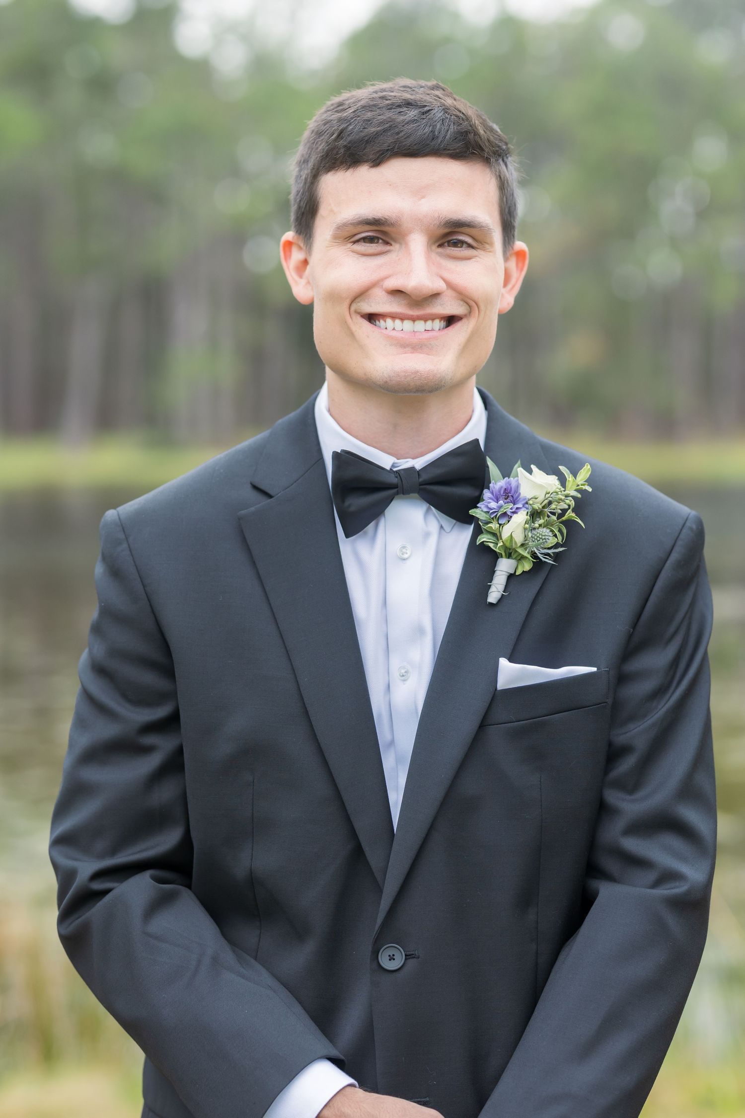 A groom in a classic black tuxedo and bowtie with a purple floral boutonniere smiles outdoors.
