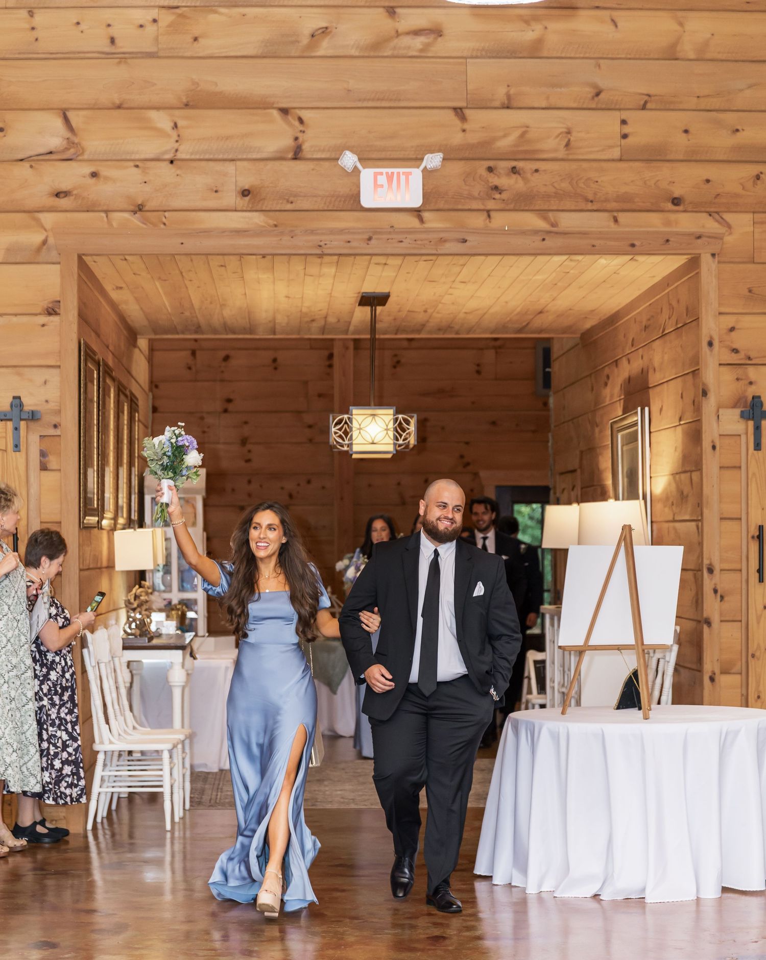 Wedding guests mingle in a rustic wooden barn venue with white tables and natural lighting.