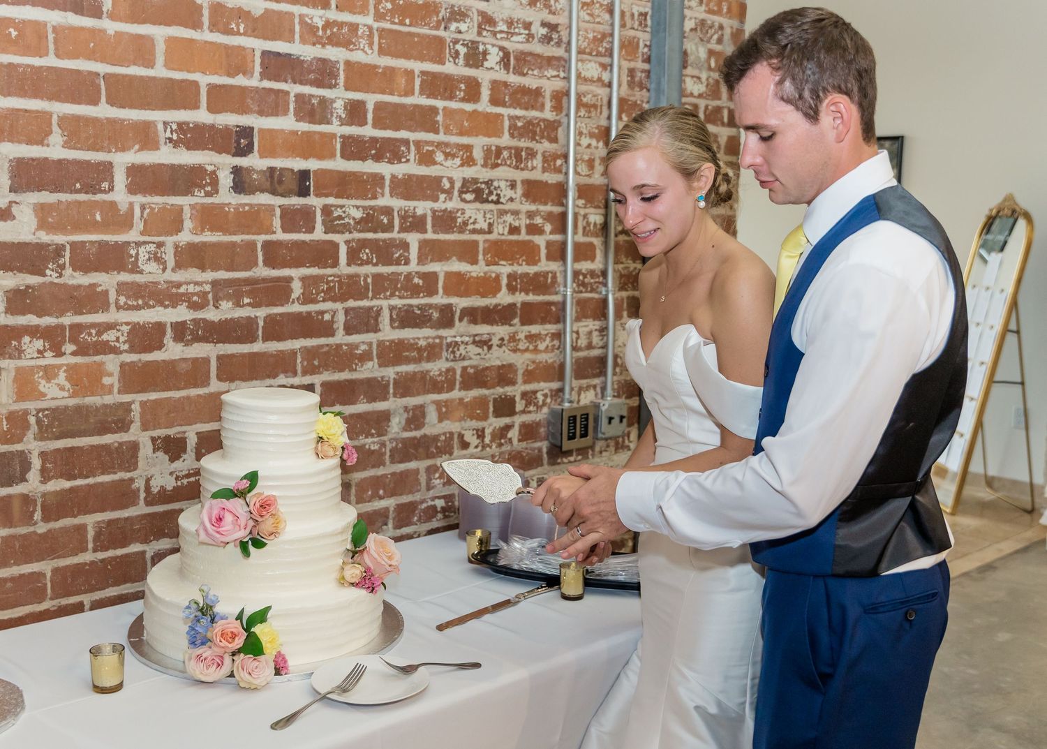 Couple cutting their elegant white wedding cake with pink floral accents against a rustic brick wall backdrop.