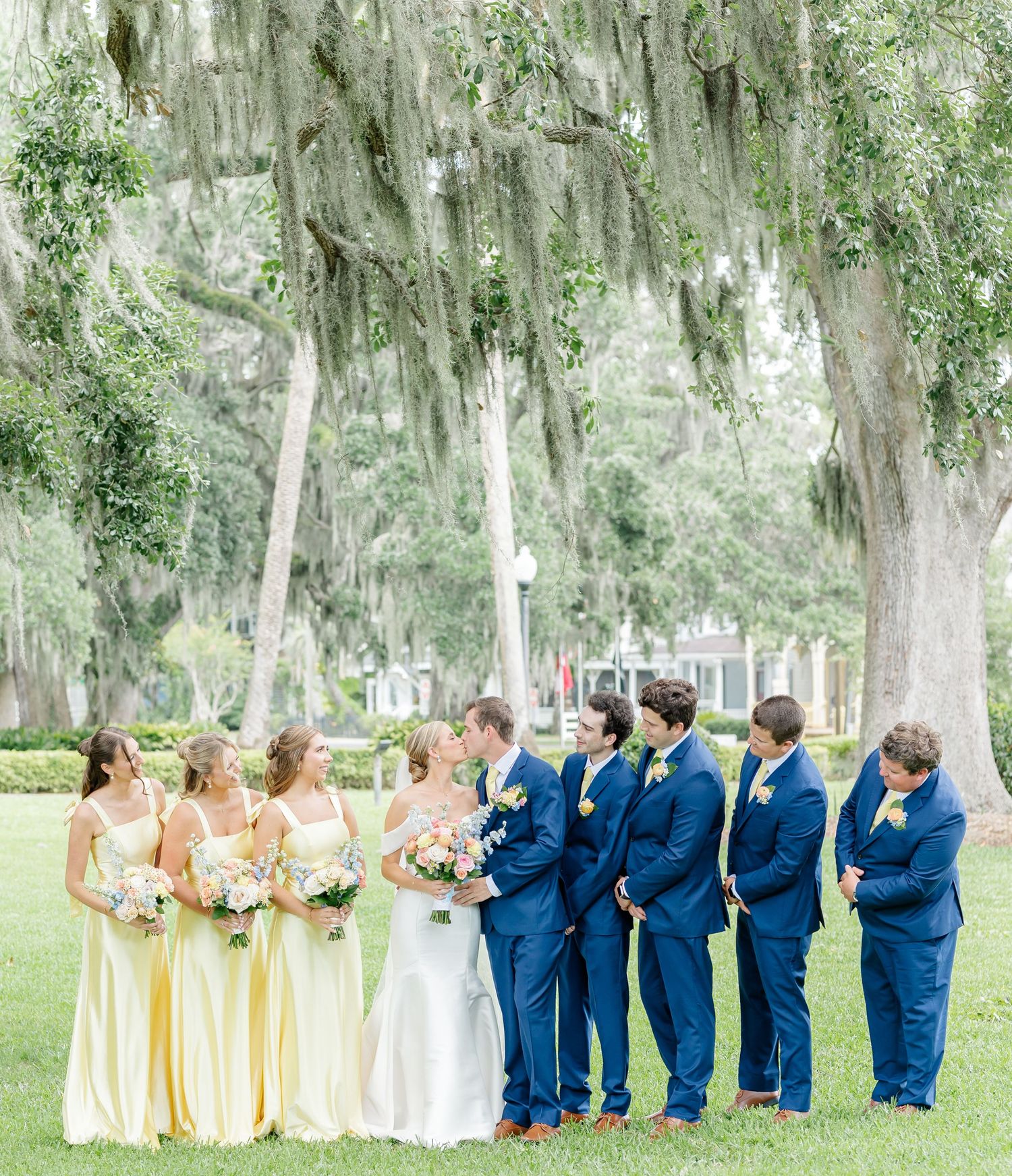 Wedding party in navy blue suits and yellow dresses poses beneath Spanish moss-draped trees in an outdoor garden setting.