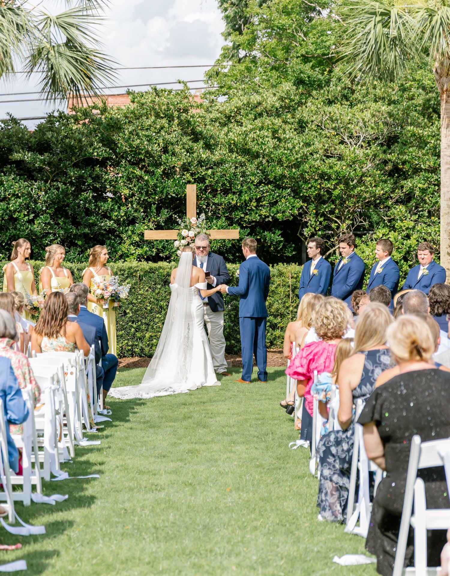 Panoramic view of an outdoor wedding ceremony with guests seated on white chairs on a lush green lawn surrounded by palm trees.
