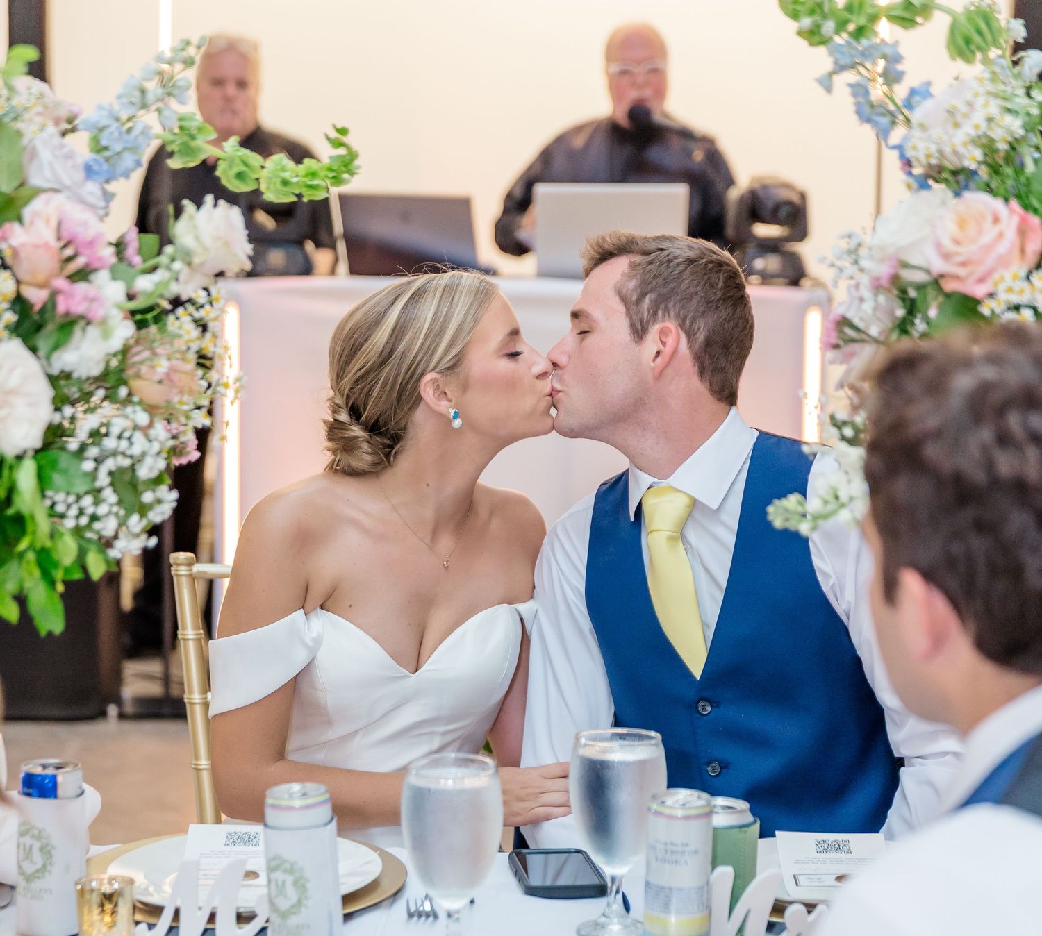 Wedding reception dinner scene with elegant floral centerpieces and table settings in light-filled venue.