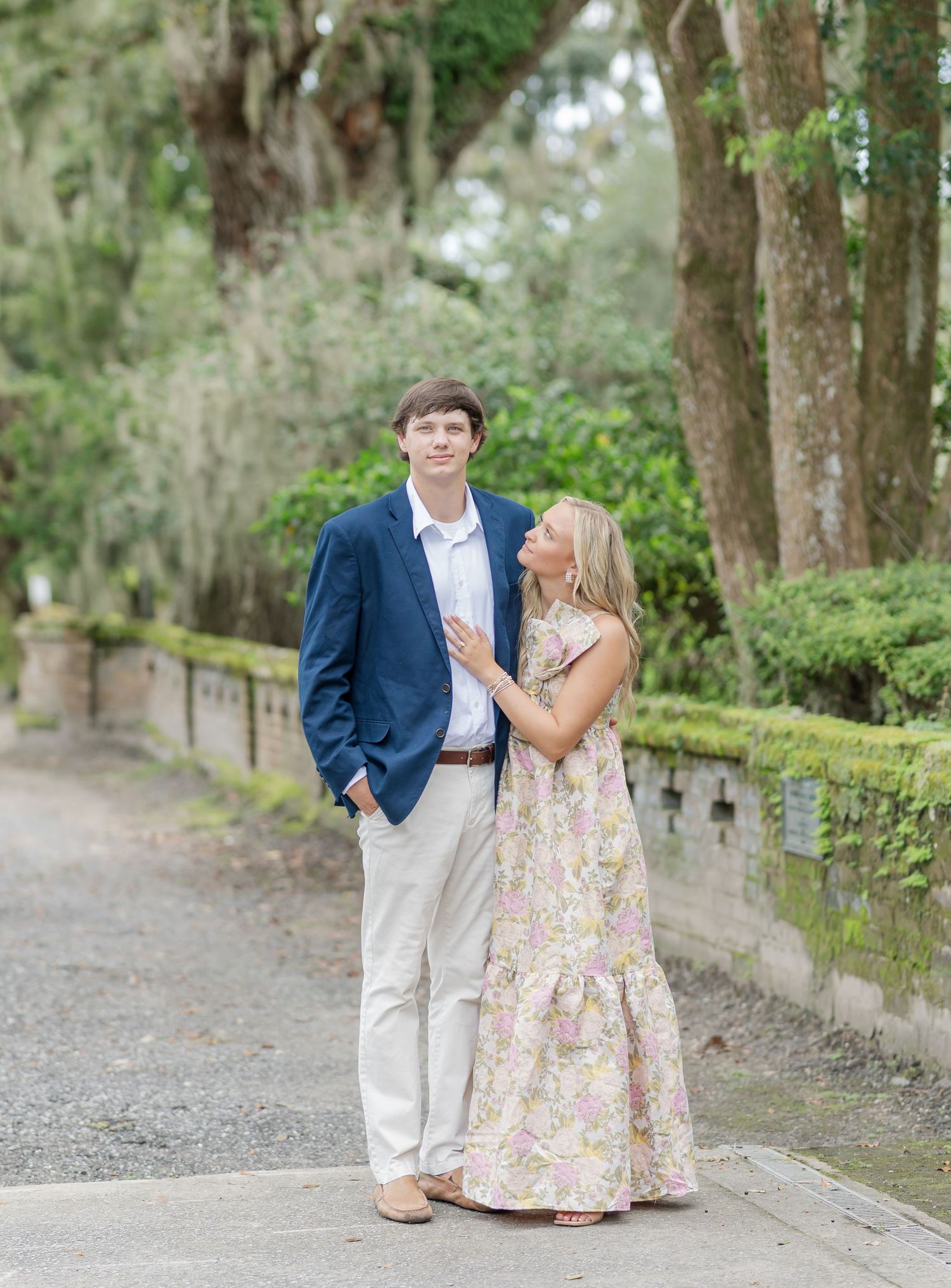 A couple shares a tender moment on a gravel path surrounded by green foliage and Spanish moss.