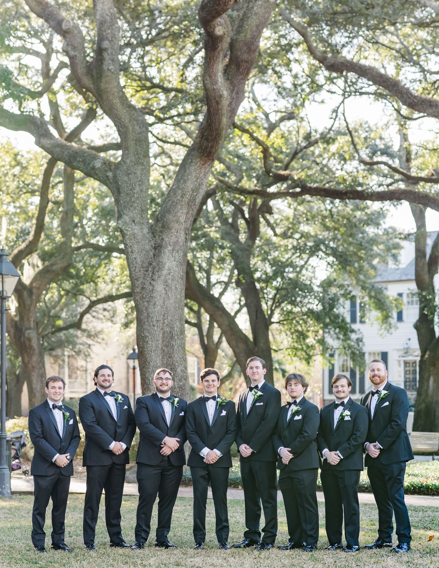 A group of groomsmen in black tuxedos stand together under large oak trees at an outdoor wedding venue.