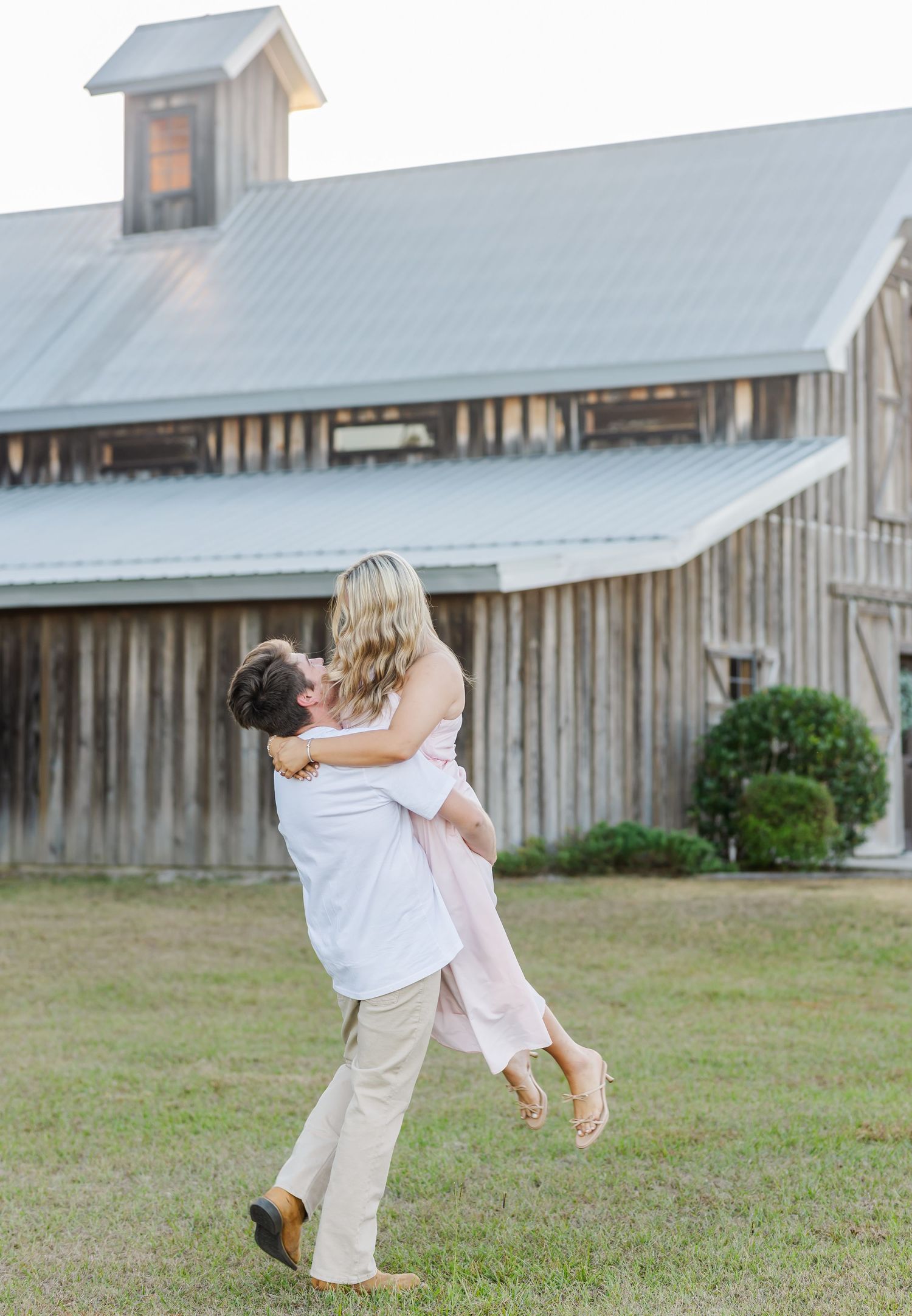 Playful couple spins and dances in front of a rustic barn during sunset.