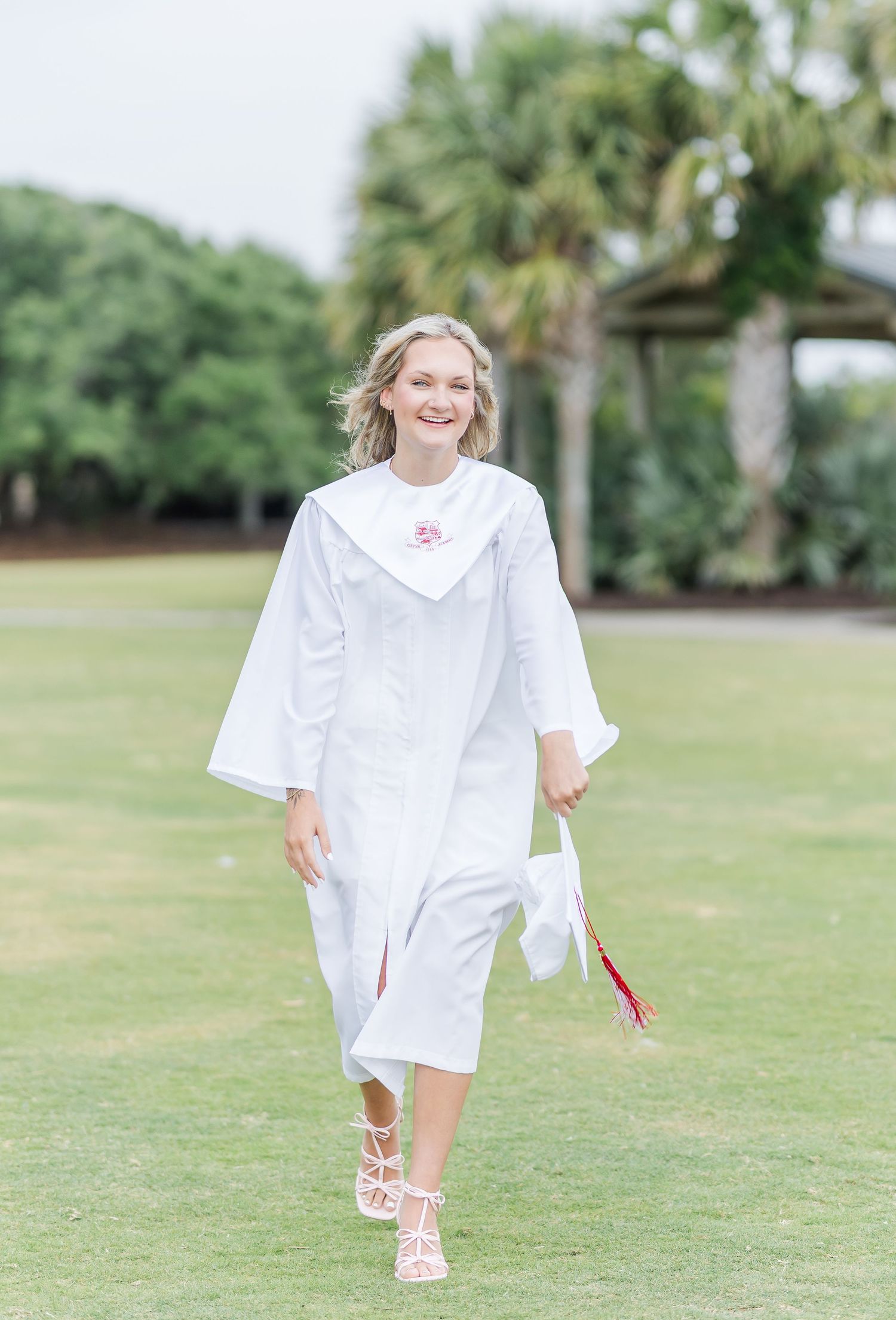 Graduate in white cap and gown walking on grass with palm trees in background during outdoor ceremony.