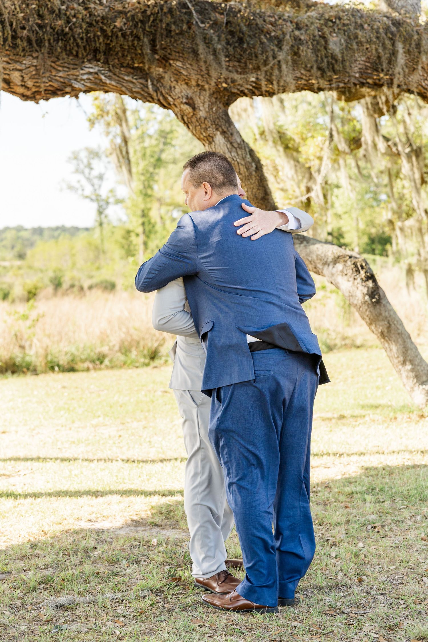Two men in suits sharing an embrace under a moss-draped oak tree outdoors.