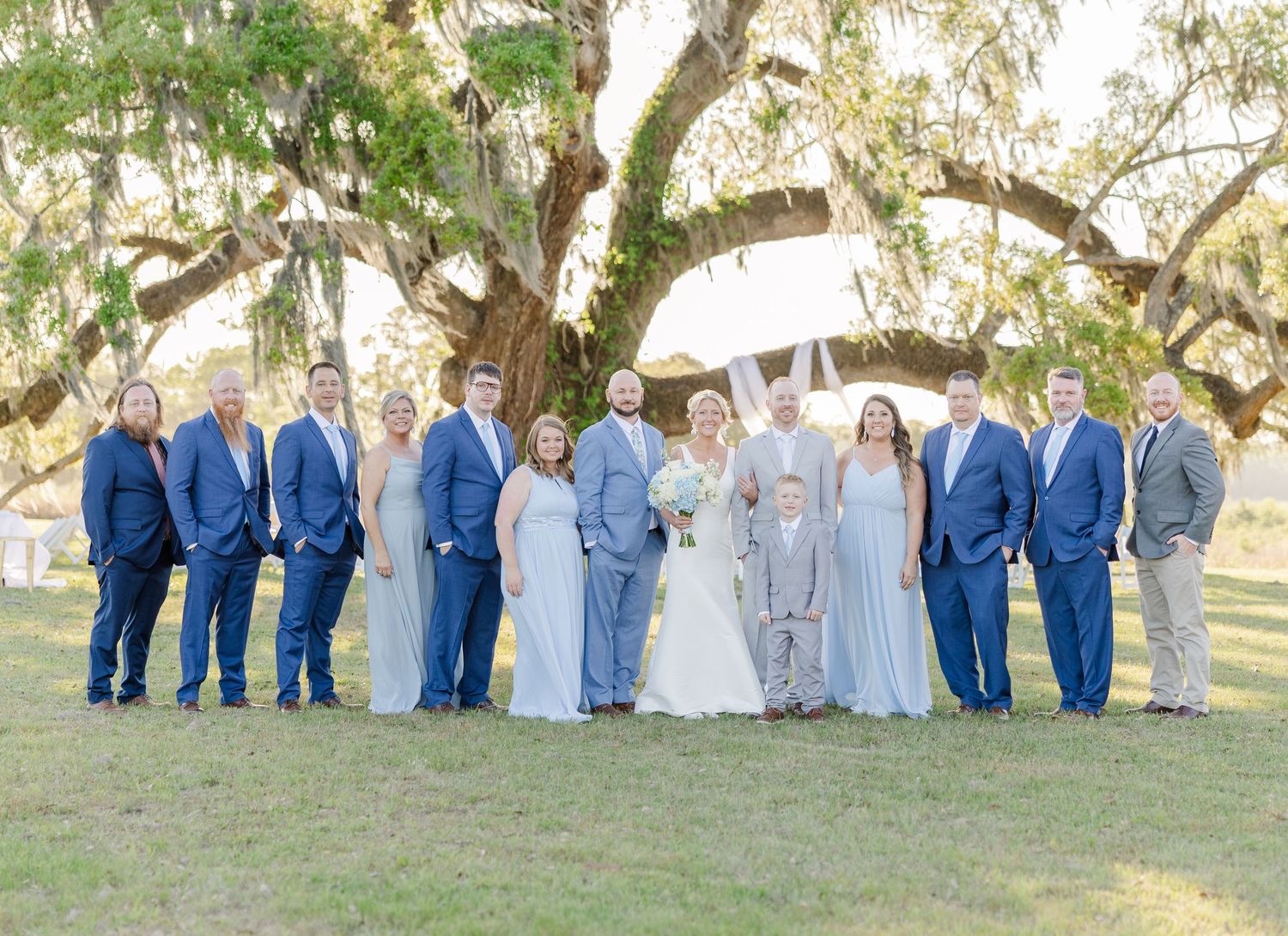 Wedding party group portrait under large oak tree with bridesmaids in blue gowns and groomsmen in navy suits.