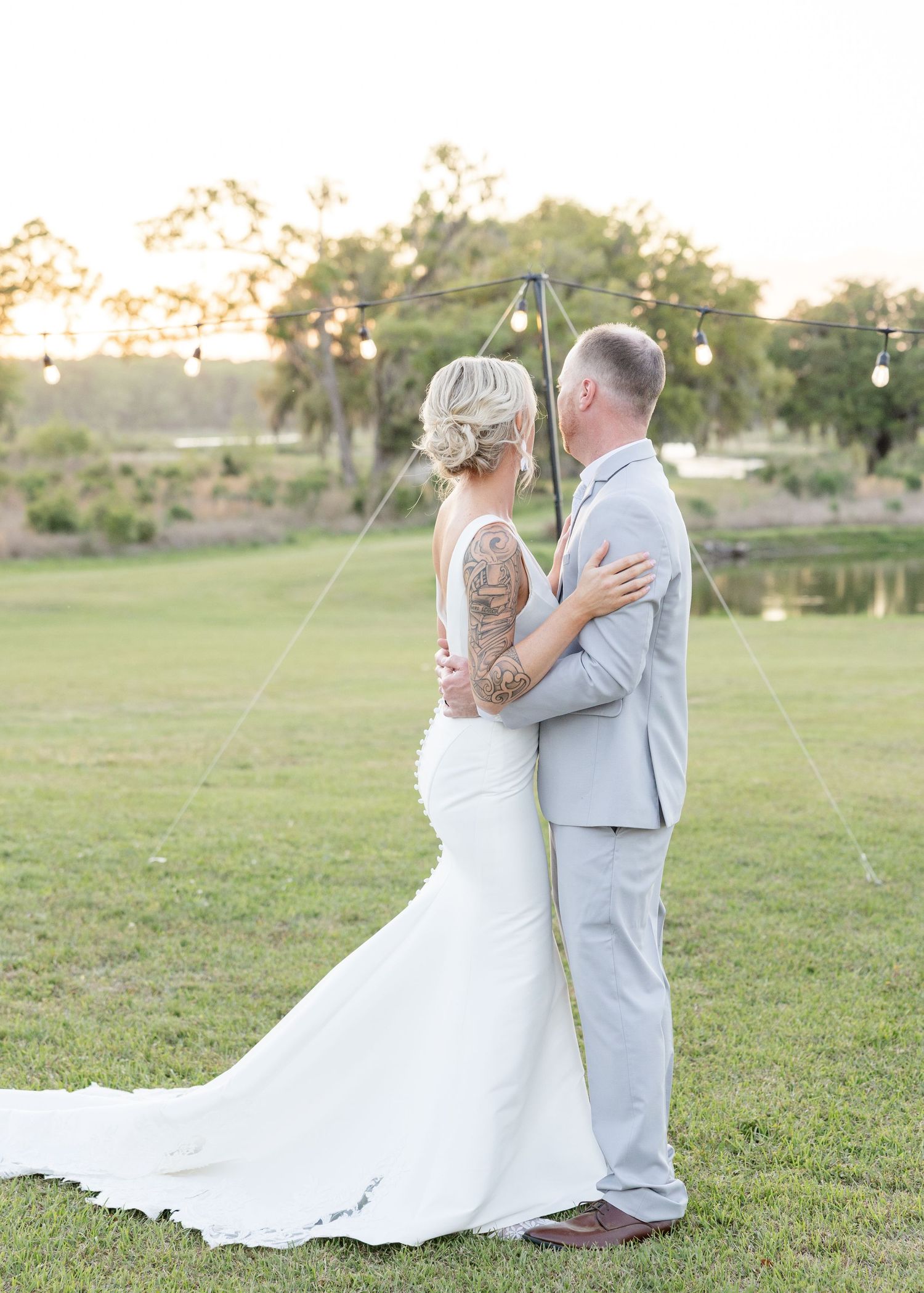 Bride with tattoo sleeve and groom embrace at sunset in front of pond with string lights.