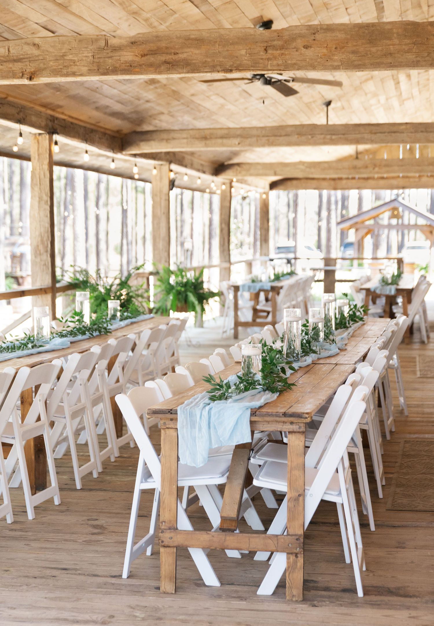 Rustic wedding reception venue with long farmhouse tables, white folding chairs, and greenery centerpieces under wooden beamed ceiling.