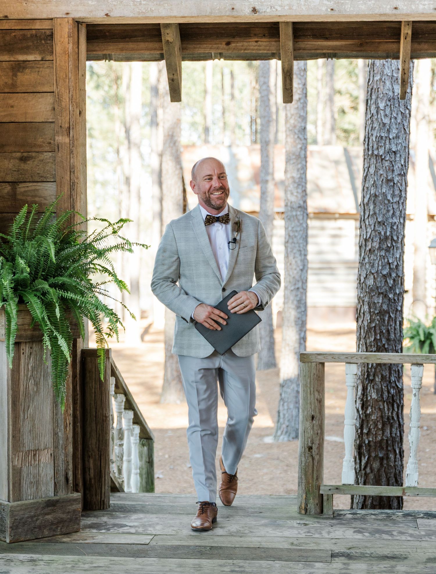 Smiling groom in light gray suit with bow tie walking down wooden ceremony aisle among pine trees.