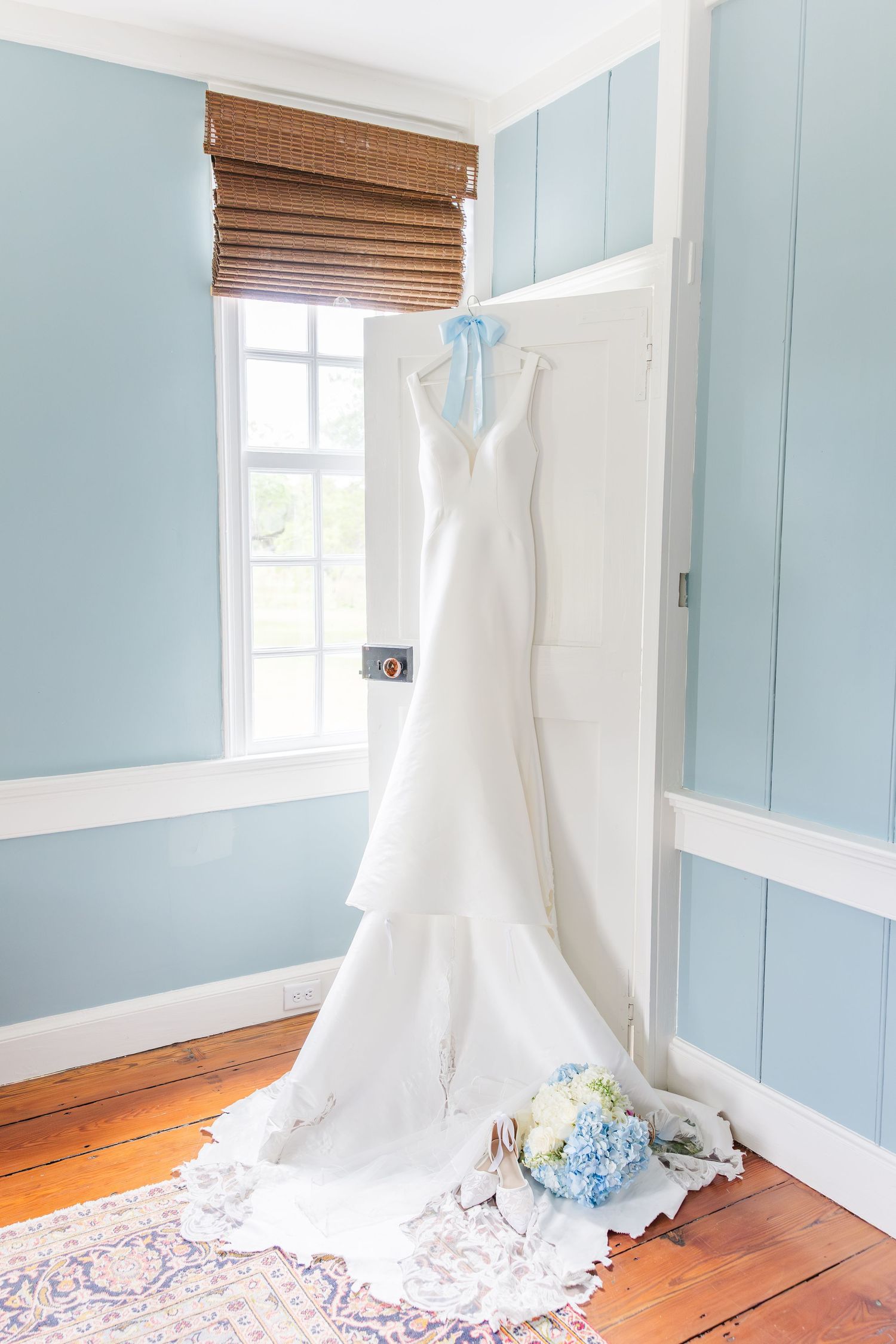 White wedding gown hanging on door with blue hydrangea bouquet and bridal accessories in light blue room.