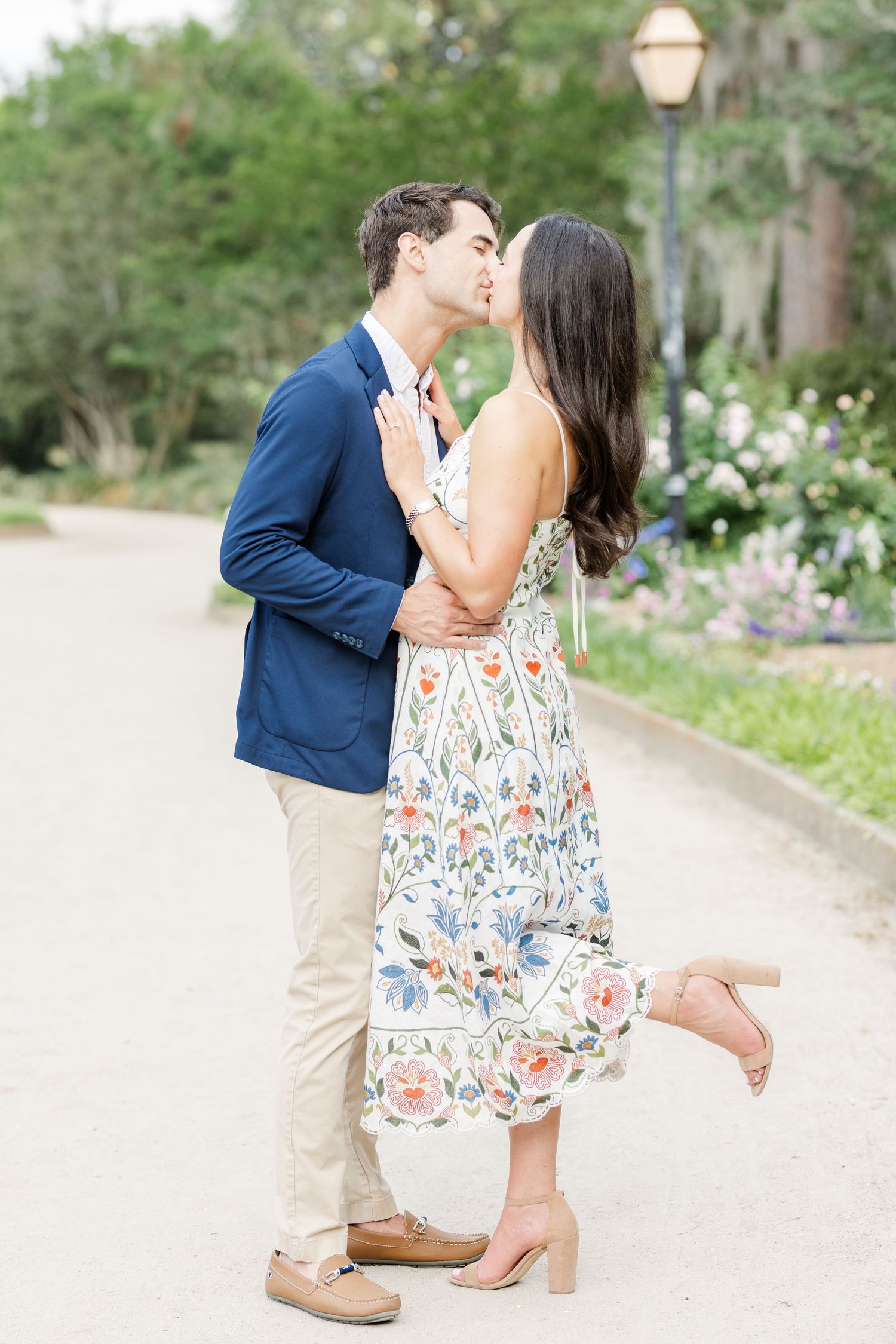 Couple shares a romantic kiss on a garden pathway, man in navy blazer and woman in white floral dress.