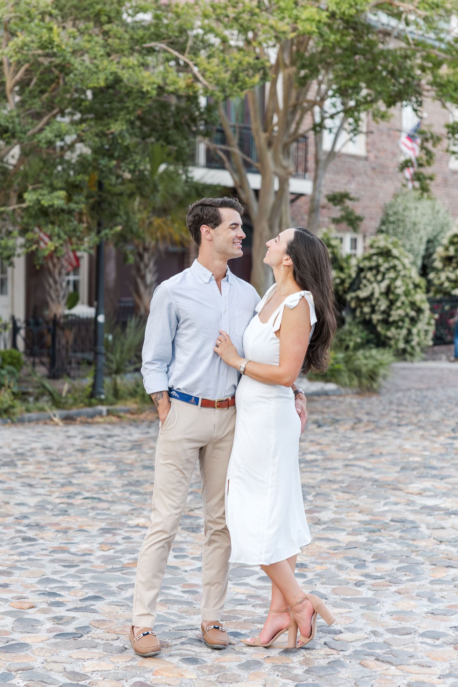Happy engaged couple smiling at each other while standing on a cobblestone street surrounded by trees.