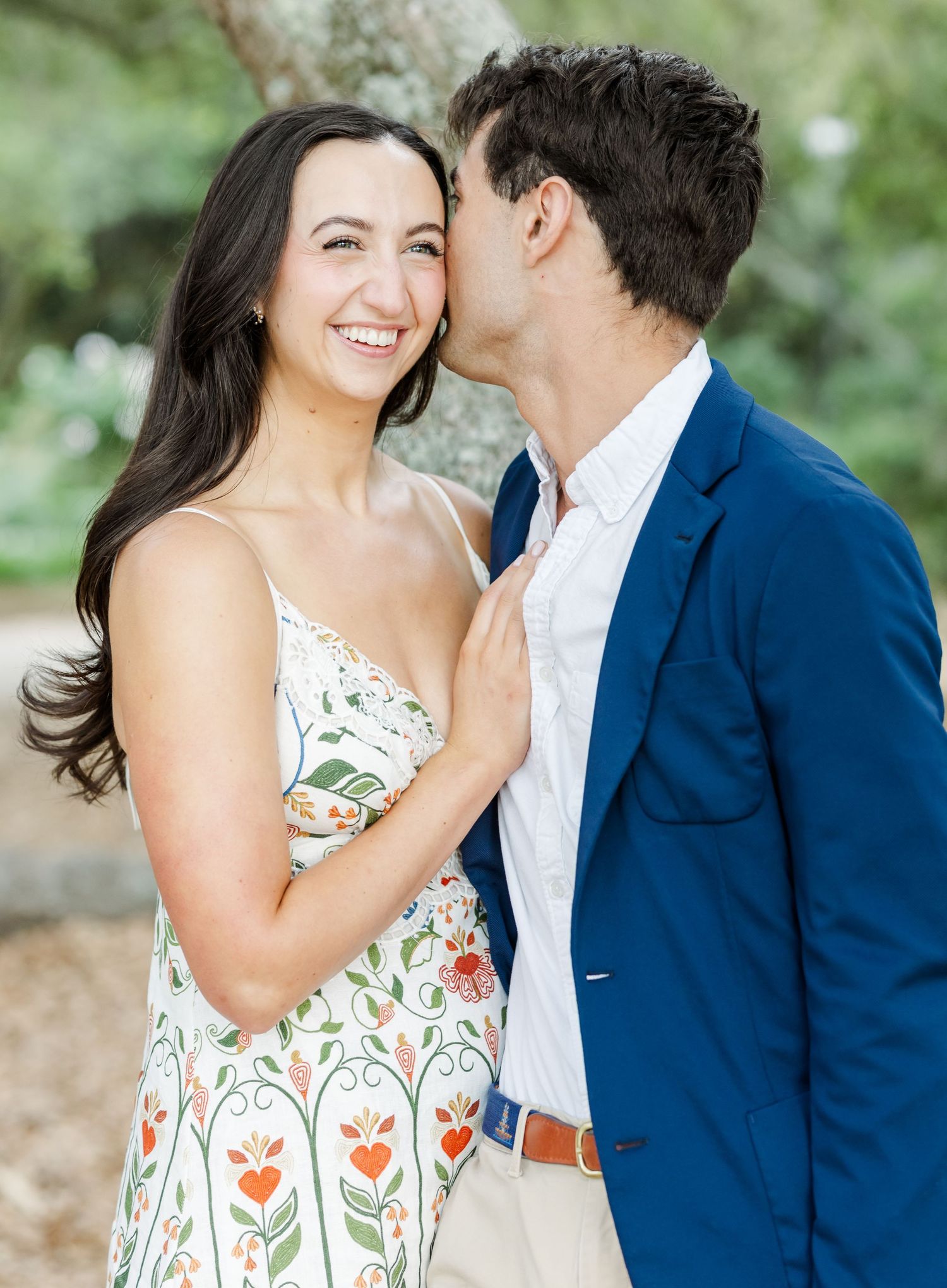 A joyful woman in a floral dress smiles as her partner kisses her cheek during an outdoor engagement session.