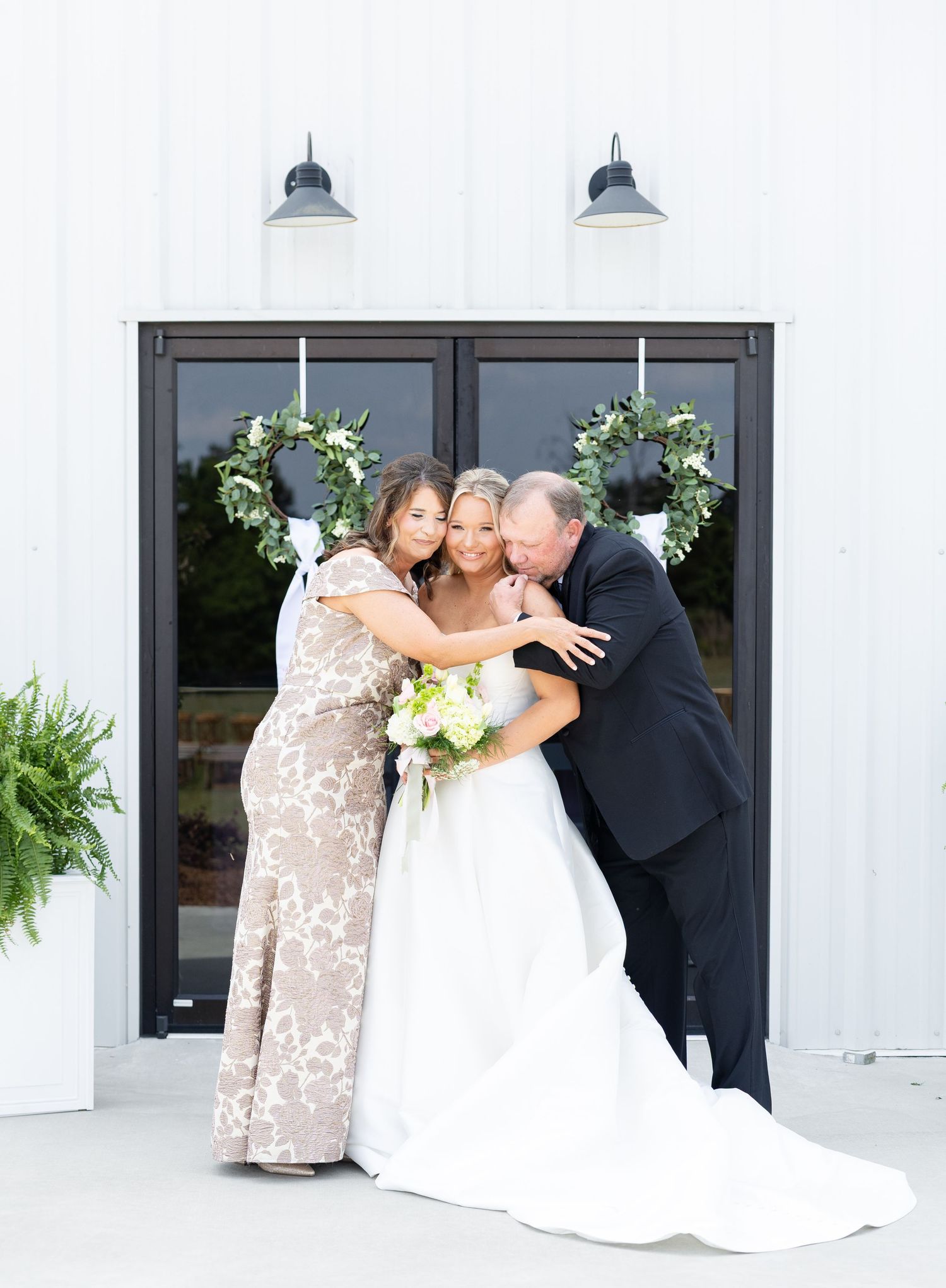 Bride in white gown holding bouquet embraced by parents in front of white barn venue doors.