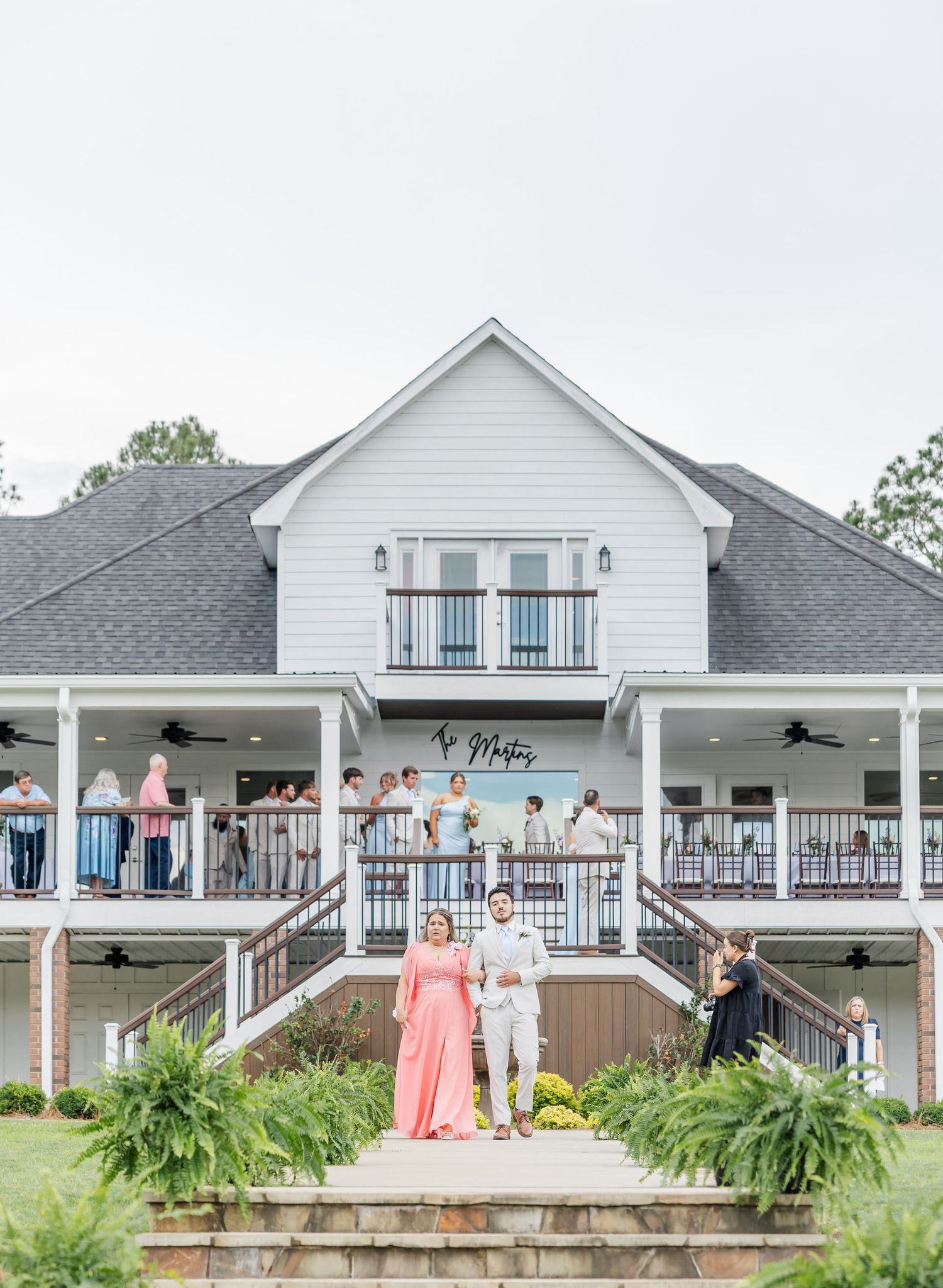 Wedding couple descends venue steps as guests watch from balcony of white farmhouse venue.