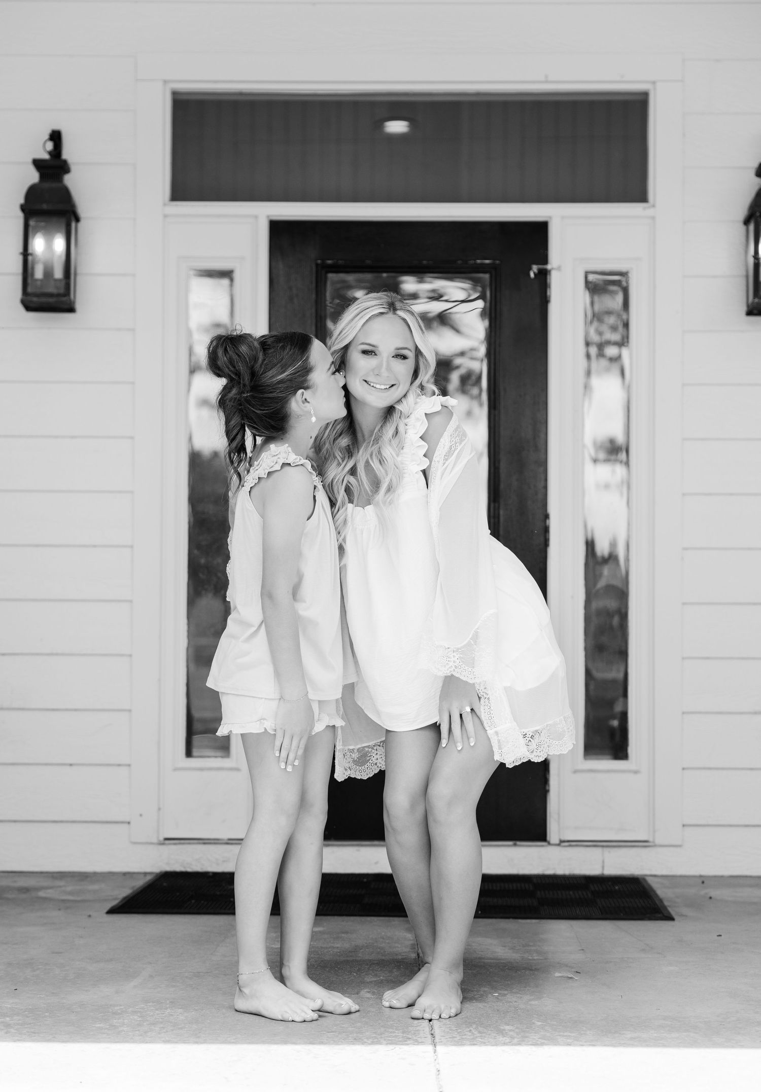 Two women in white outfits sharing a joyful moment outside a farmhouse door in black and white.