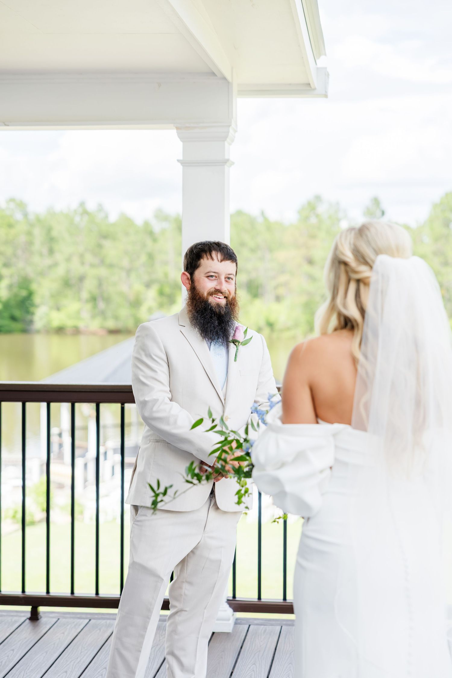 Bearded groom in light gray suit smiles at bride during outdoor first look on waterfront venue porch.