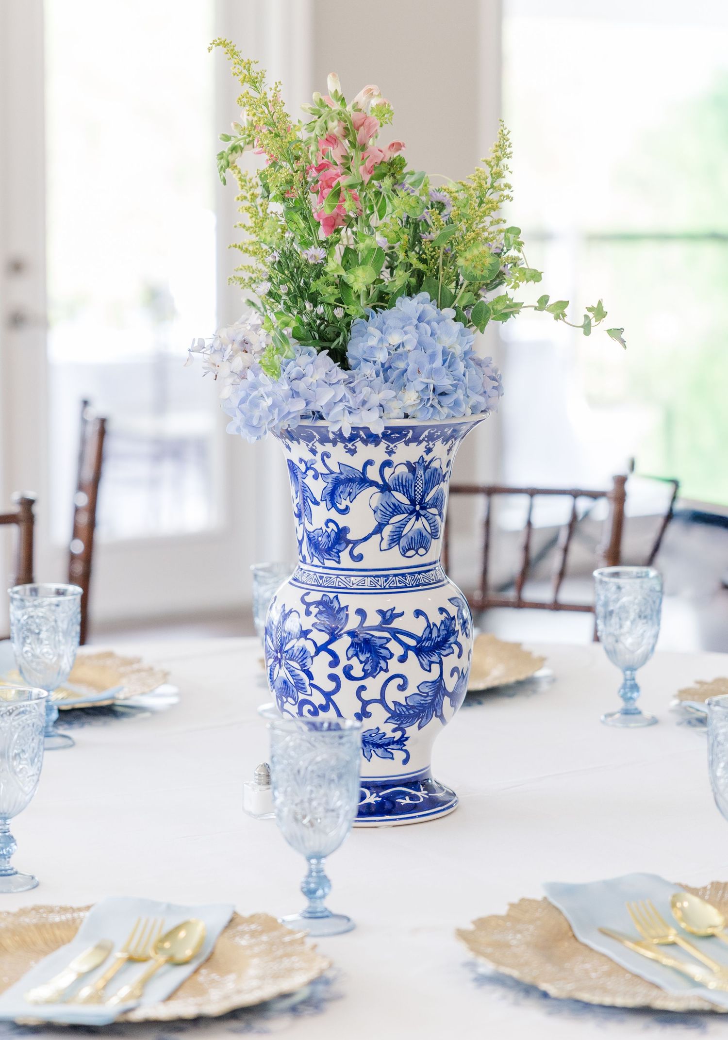 Elegant blue and white chinoiserie vase centerpiece with hydrangeas and pink flowers on a white tablecloth.