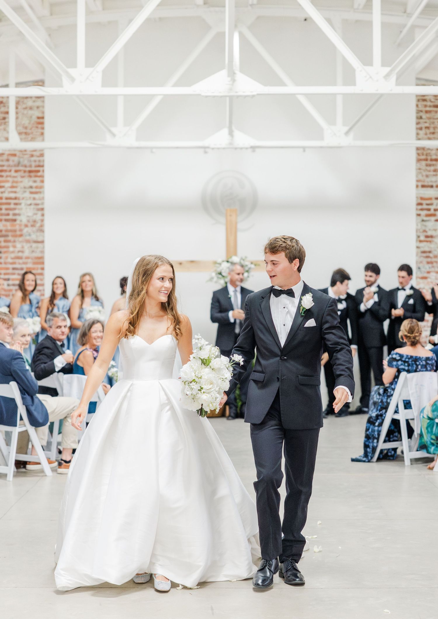 Newlyweds walk back down aisle after ceremony in bright industrial wedding venue with exposed brick.