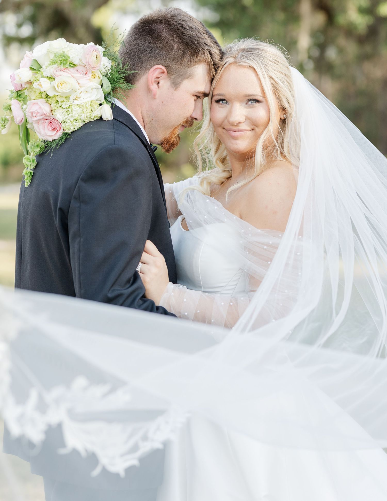 Bride in lace gown and flowing veil holds pink bouquet while embracing groom in dark suit outdoors.