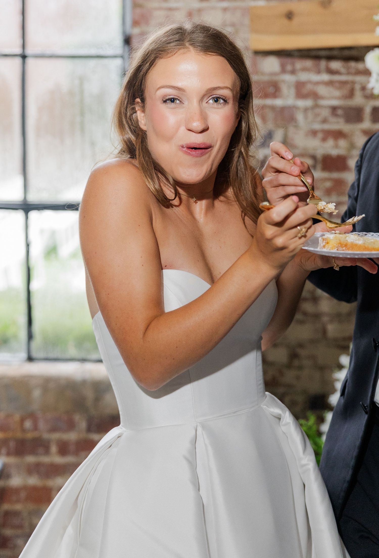 Happy bride in strapless white gown excitedly eating wedding cake at reception.