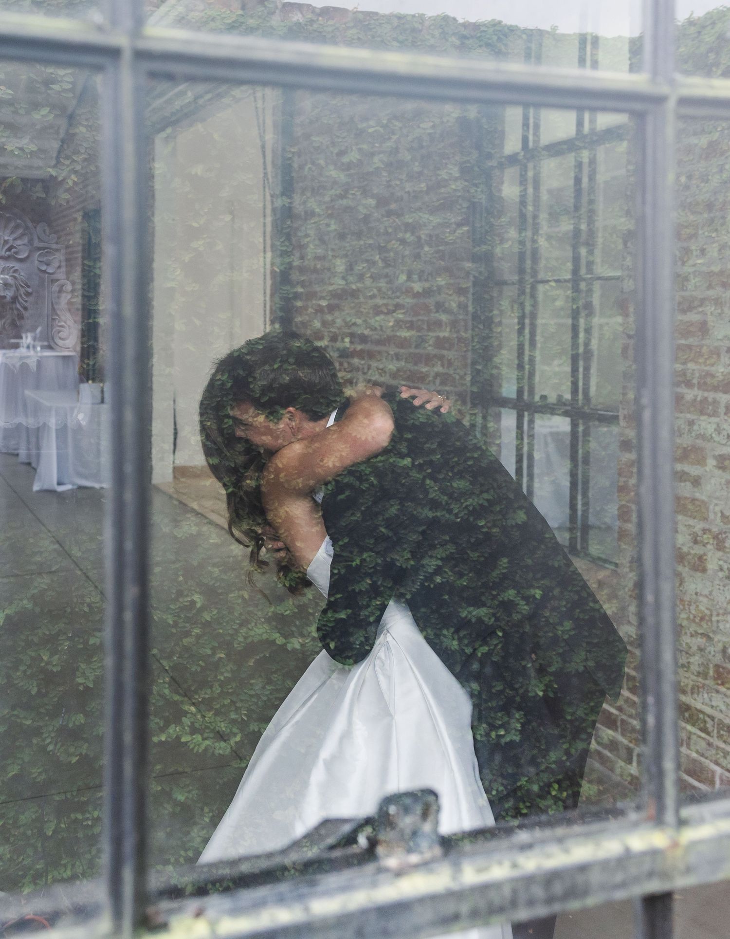 Bride and groom share romantic first kiss embrace viewed through glass window at wedding venue.