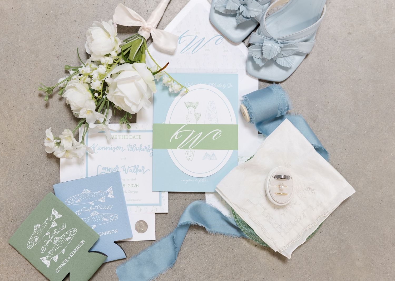 Elegant wedding flat lay with sage green stationery, white floral bouquet, dusty blue ribbon, and bridal accessories on neutral background.