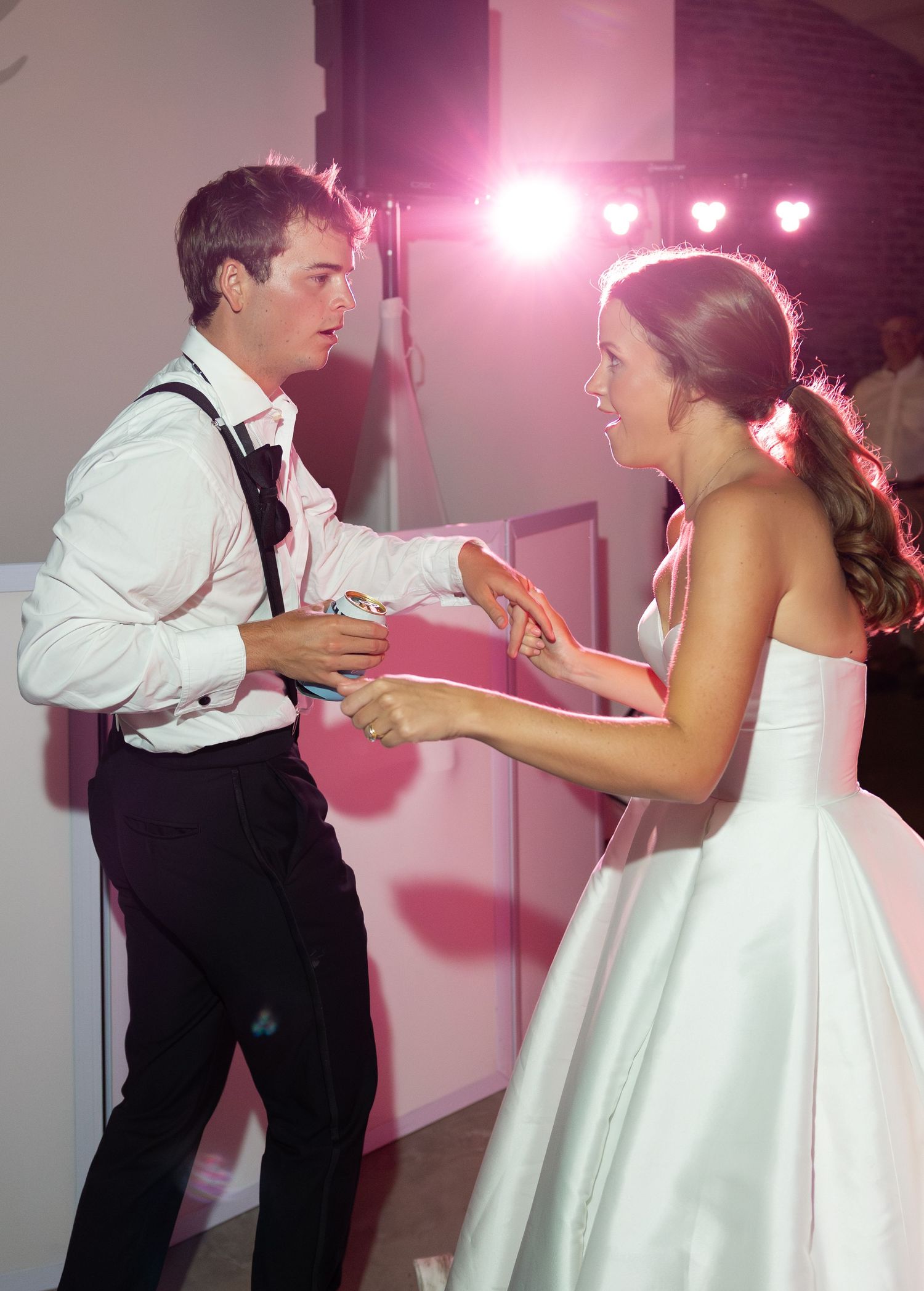 Bride in white strapless gown and groom dancing together at wedding reception under pink lights.