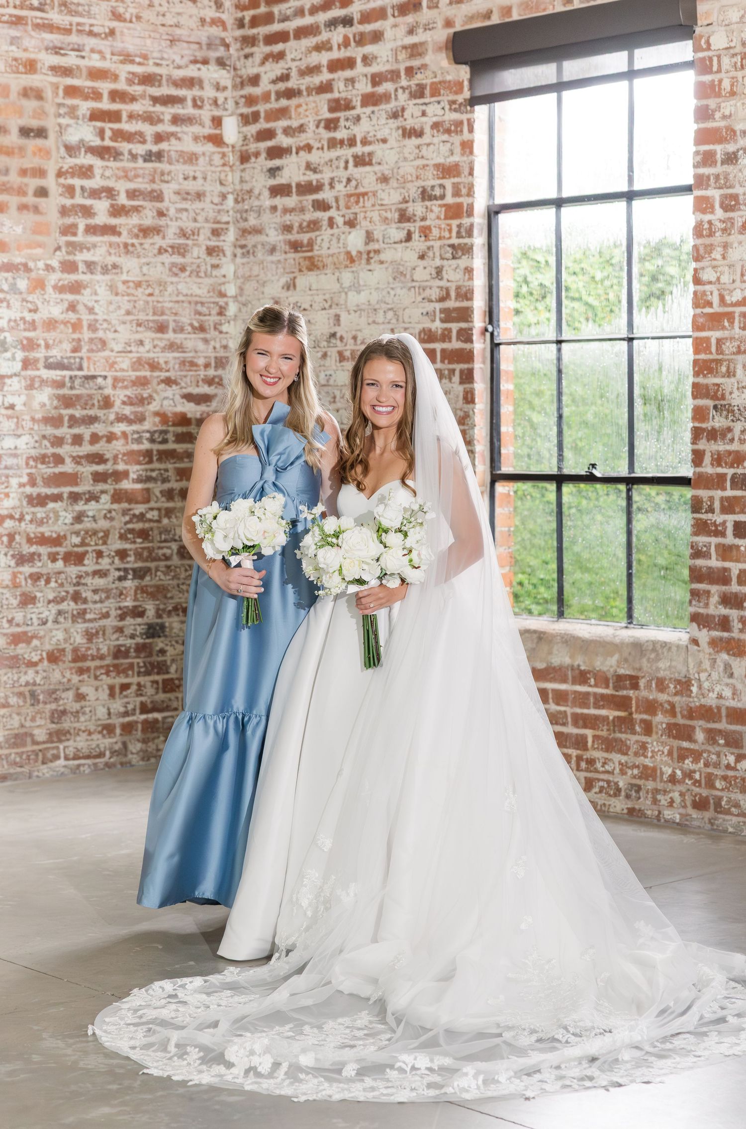 Bride in lace gown with veil poses with bridesmaid in blue dress against brick wall.