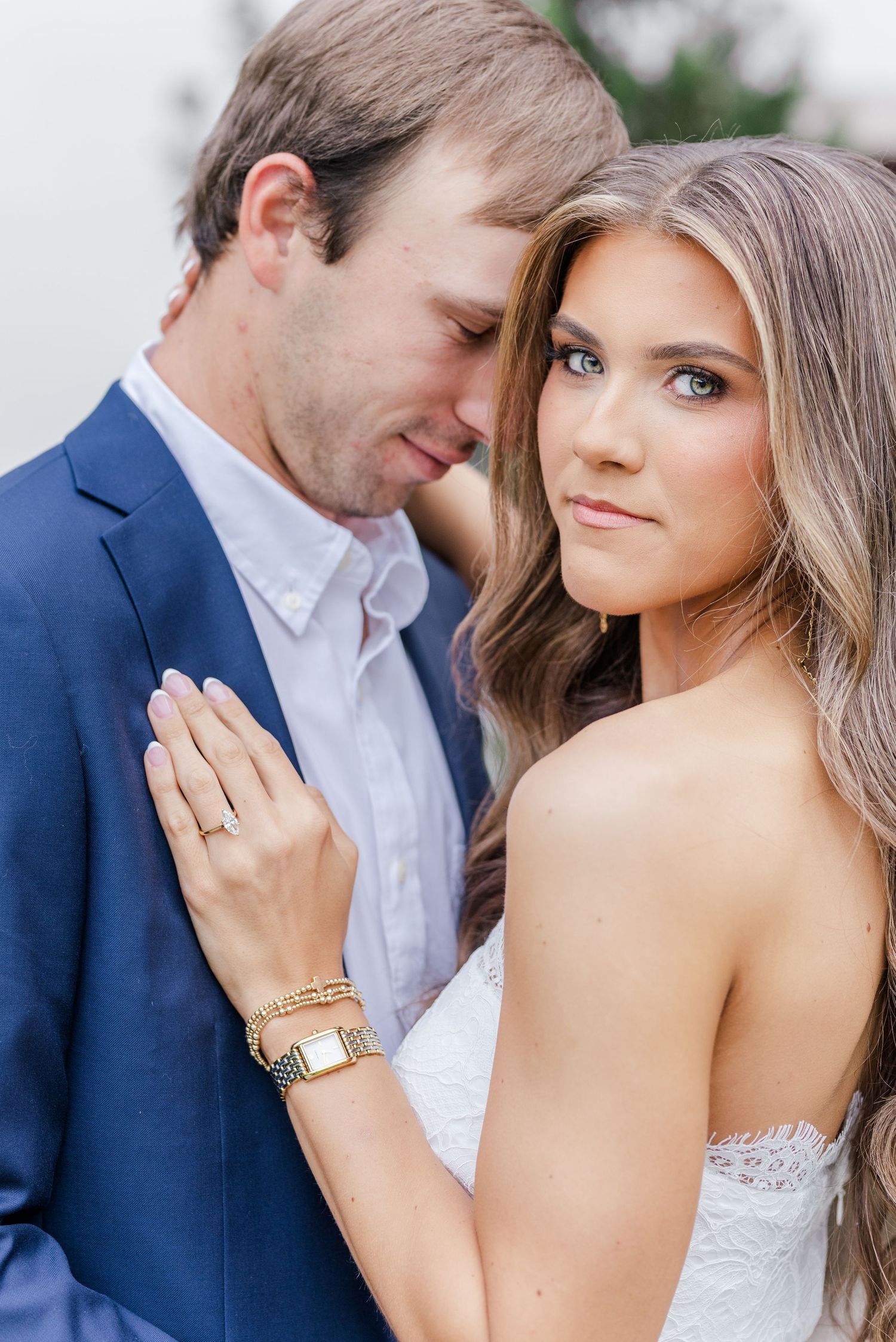 Elegant couple in engagement photo, woman in white lace dress with gold watch and ring, man in navy suit.