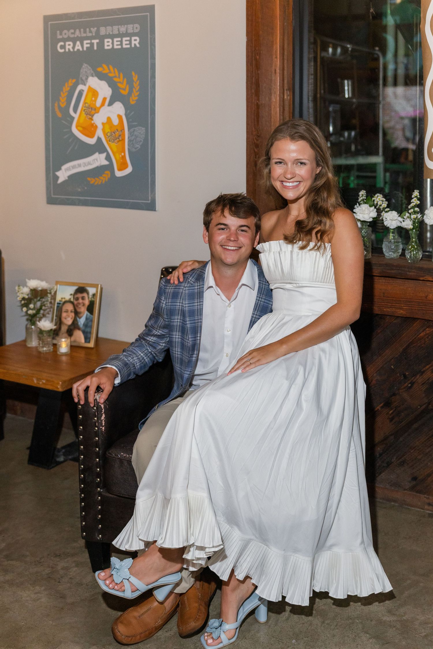 Couple poses at engagement party near craft beer sign, man in plaid blazer and woman in white dress.