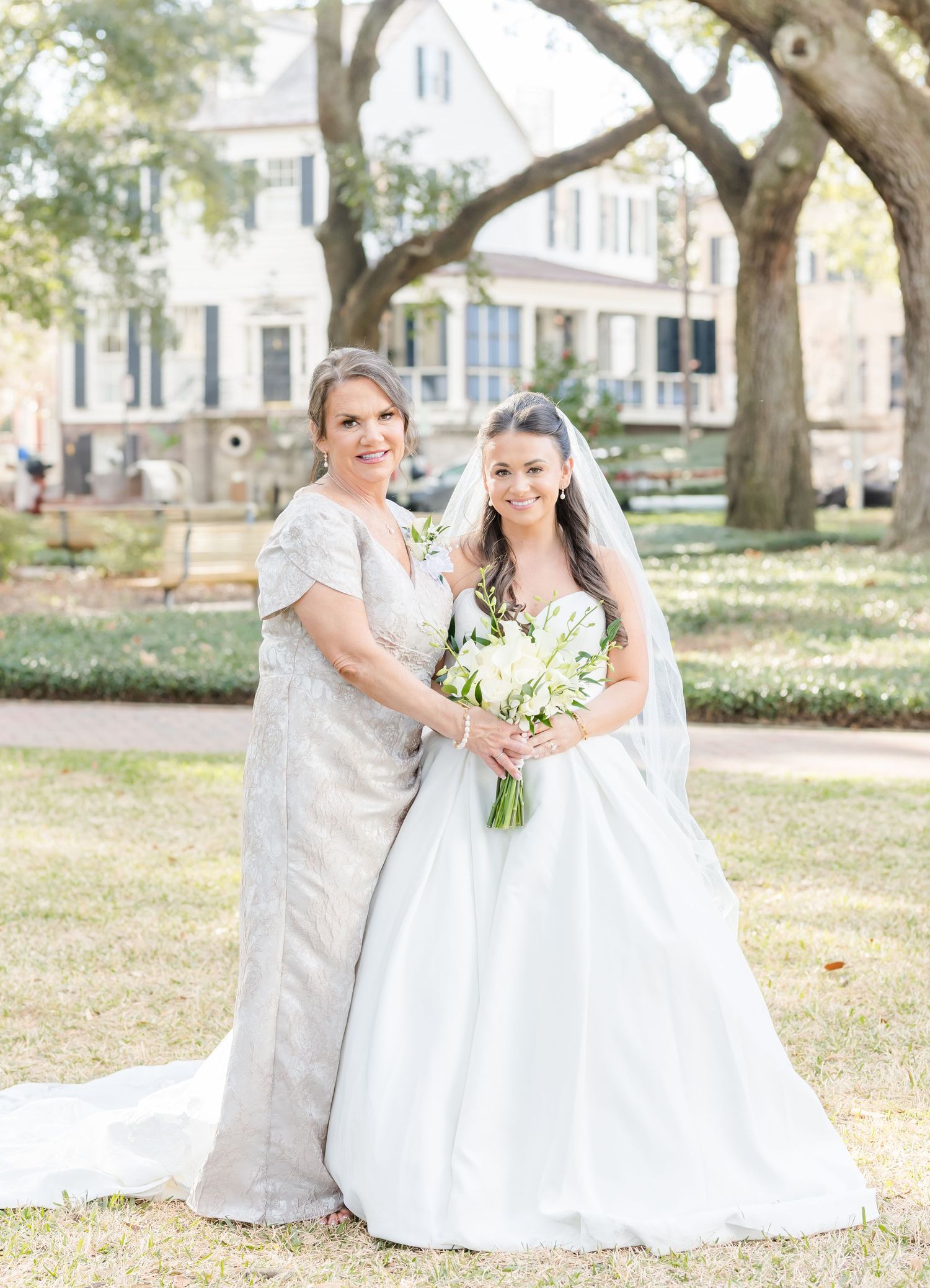Bride in white ball gown with veil poses with older woman in silver dress holding white bouquet outdoors.