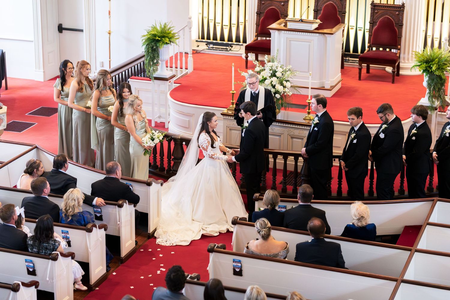 Elegant church wedding ceremony with bridesmaids, groomsmen, and guests seated in wooden pews with red carpet aisle.