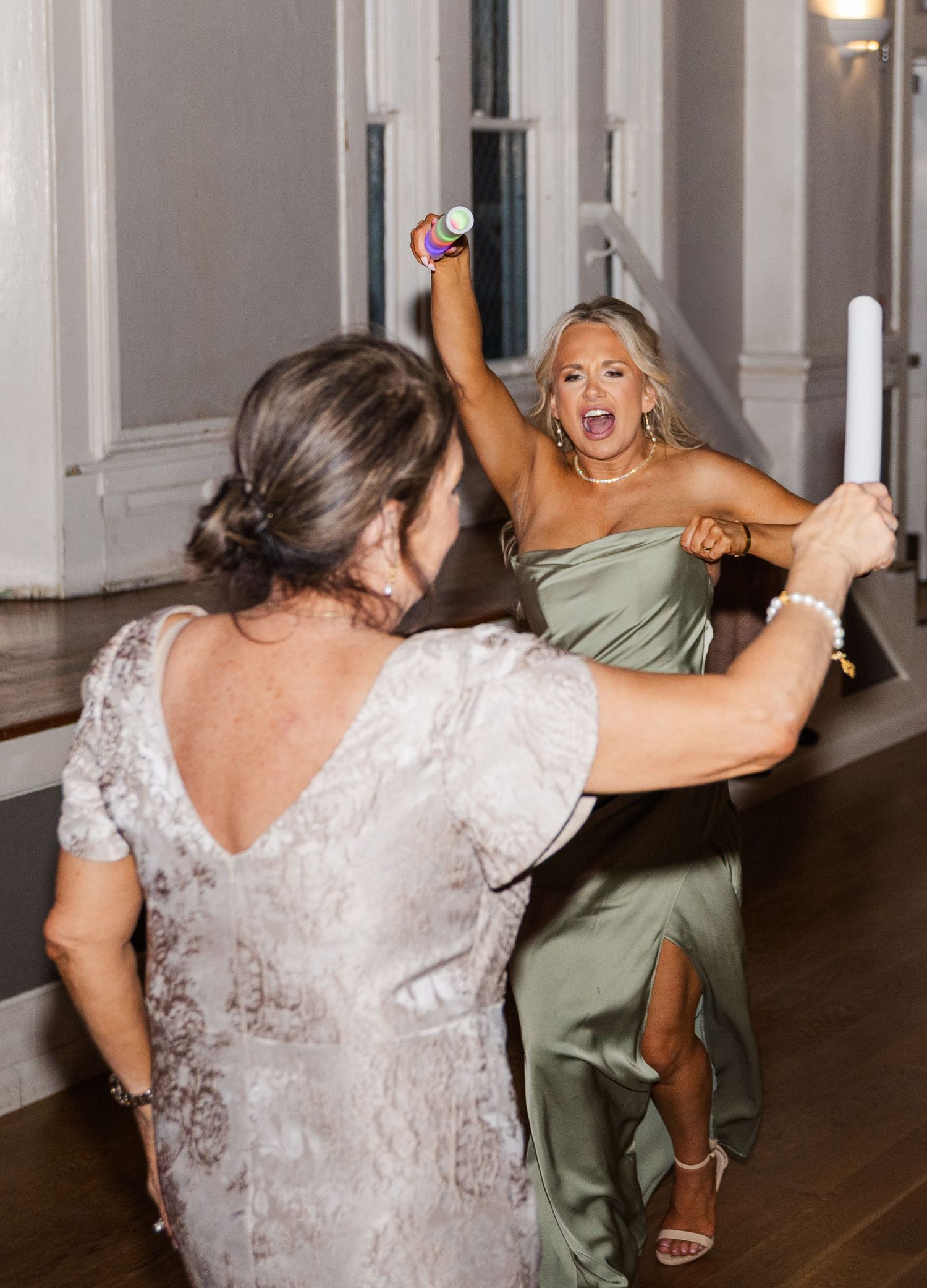 Two women dance energetically with glow sticks at a wedding reception.