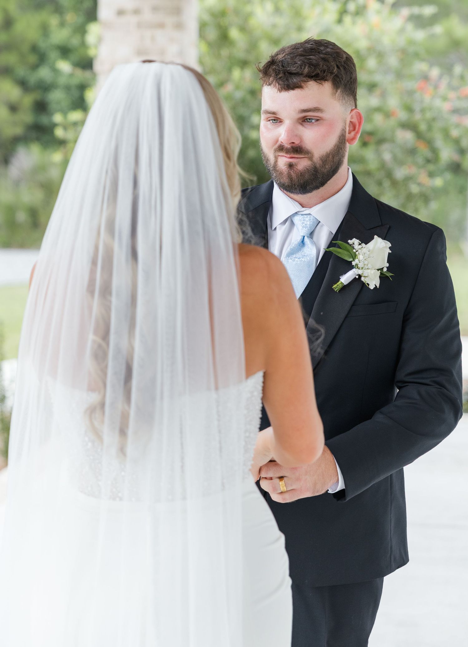 Groom in black tuxedo with blue tie holds bride's hands during outdoor first look moment.