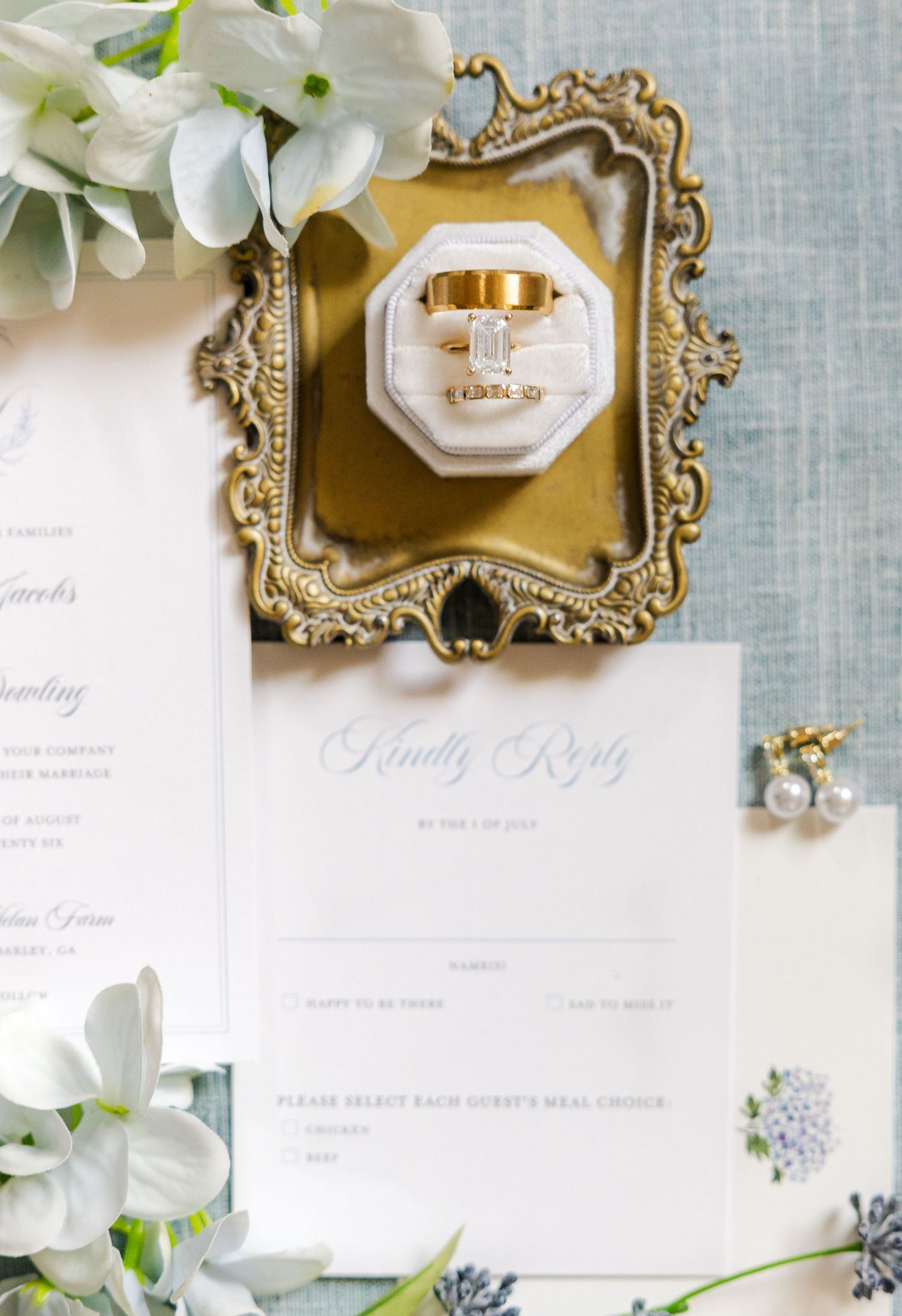 Elegant wedding flat lay with gold rings in velvet box, RSVP card, and white floral accents on ornate gold tray.