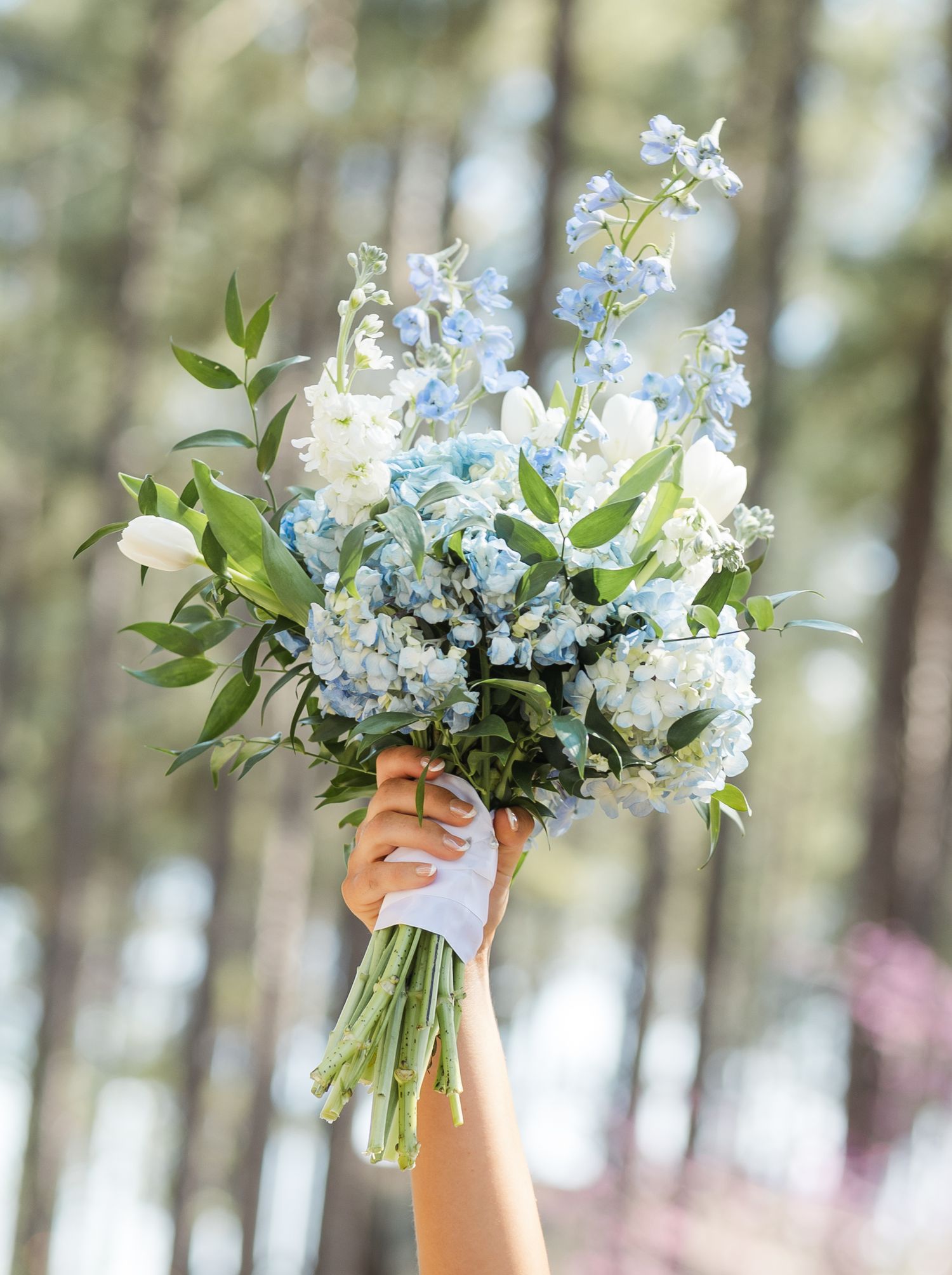 A delicate bouquet of white and light blue flowers including delphiniums and greenery held up against a blurred forest background.